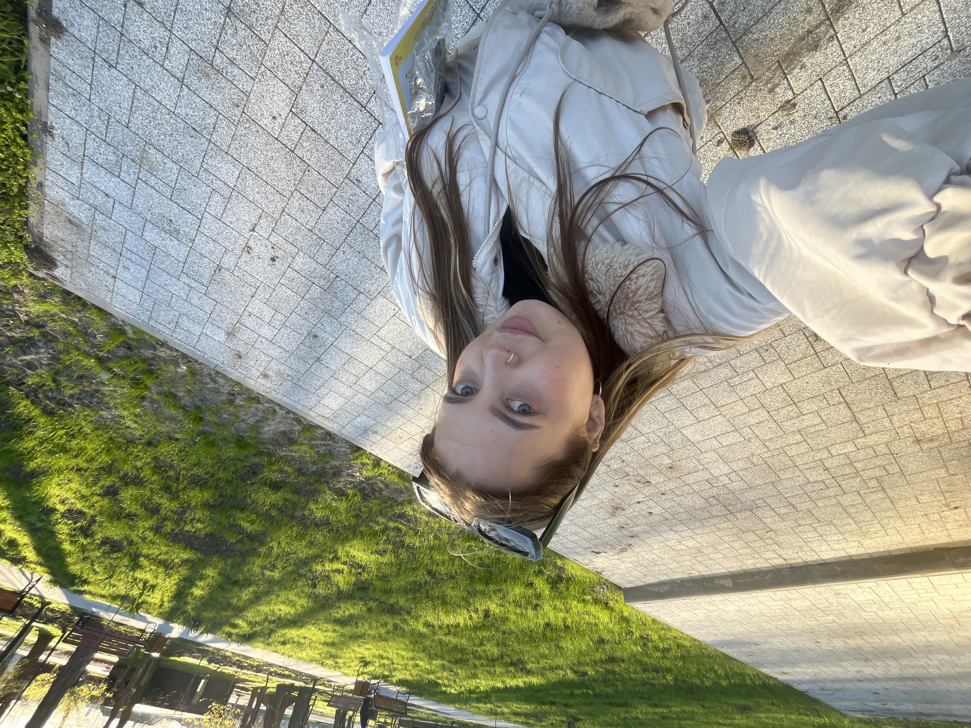 A young woman with long brown hair and blue eyes is the central figure in an upside-down outdoor selfie. She wears a light-colored jacket with a fur-lined collar. Sunglasses rest on her hair. She is standing on a paved walkway made of rectangular gray stones. To the side of the walkway is a grassy area containing benches. This scene is likely taking place in a park or public space in Novopokrovskaya, Russia. There is a book on the ground.