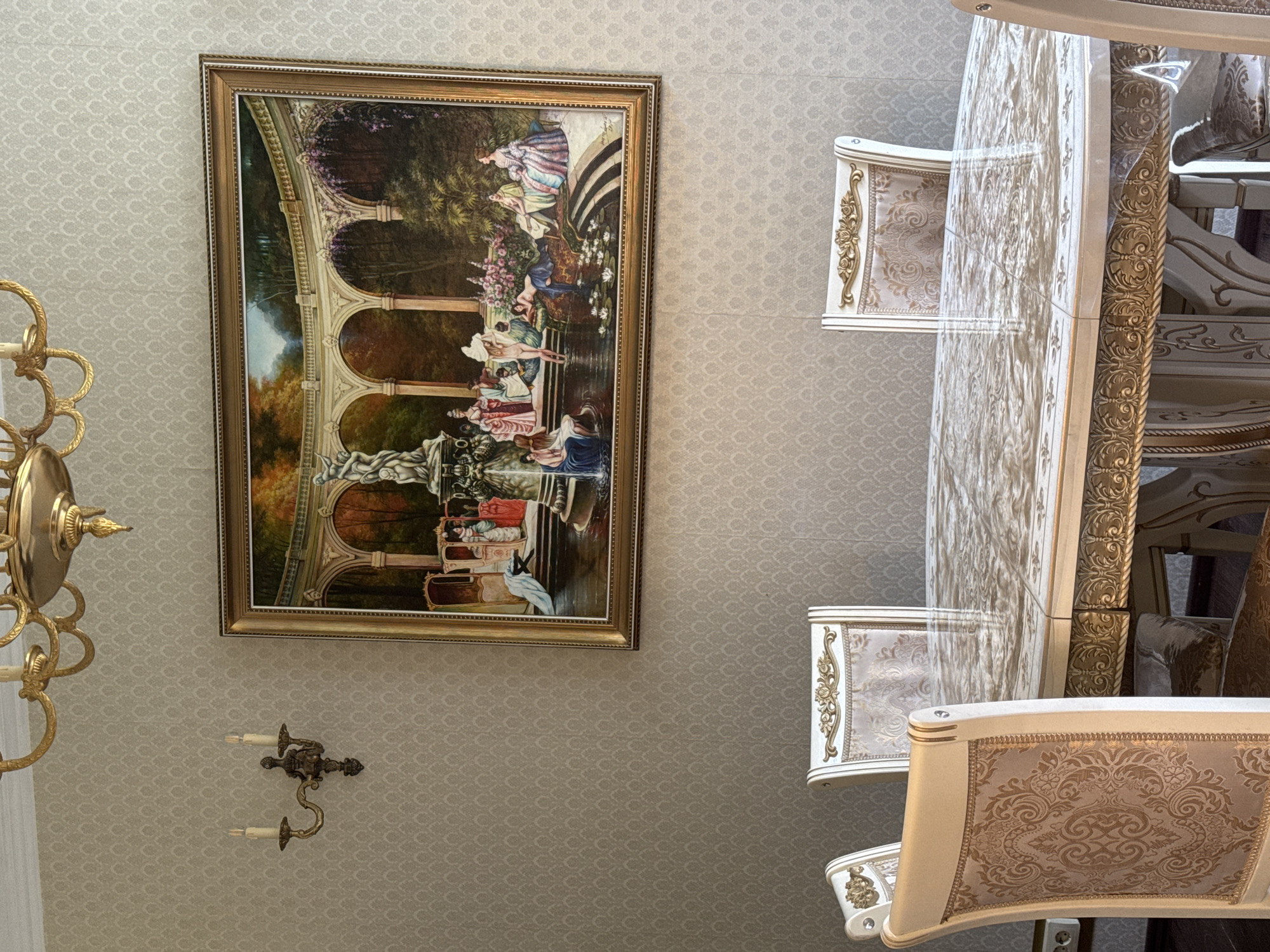The image shows an indoor dining room. The walls are covered in a pale, textured wallpaper with a subtle repeating pattern. A large, ornate framed painting hangs on the wall, depicting a scene with classical figures in a garden setting by a fountain. Above the painting, a brass chandelier with multiple arms is visible. To the left of the painting, a brass wall sconce with two candle-like lights is mounted on the wall. In the foreground, a large oval dining table with a marble-patterned top is covered with a clear plastic protective sheet. The table has ornate carved details and is surrounded by several upholstered dining chairs with decorative patterns in gold and cream. The chairs have high backs with golden ornate trim and upholstered seats. The scene appears to be a well-decorated dining area, possibly in a home.