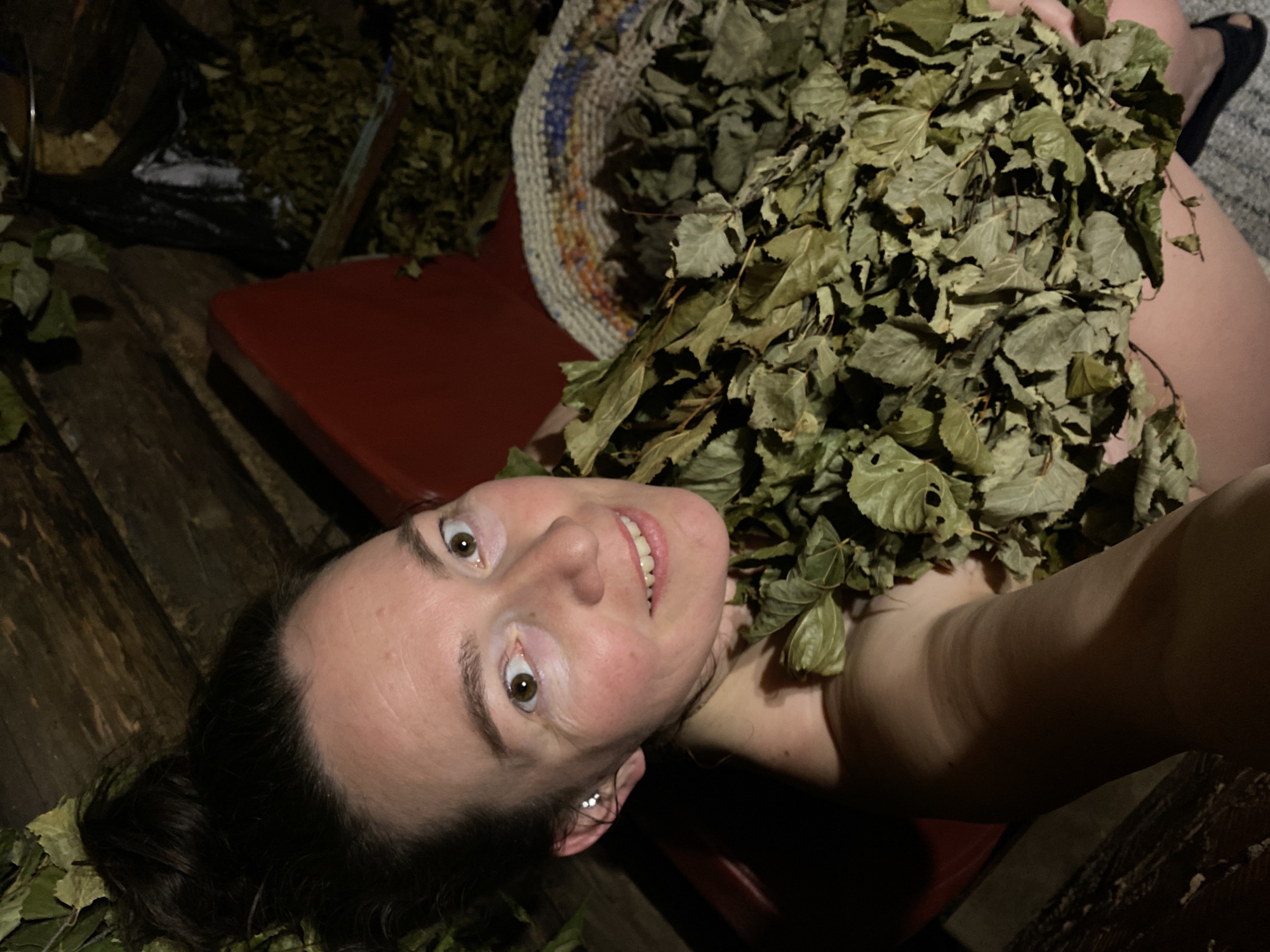 A woman lies prone in a wooden sauna, surrounded by birch branches used for venik massage. She smiles at the camera, her face slightly flushed. The sauna's interior includes wooden planks, a red bench, and a woven mat. The location is not specified as the city of Noviy, Russia.