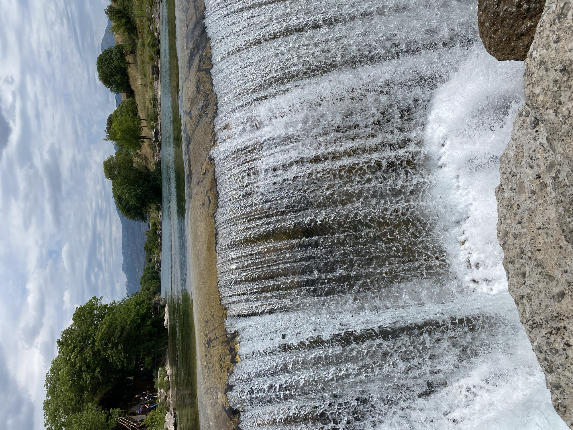 The image, best viewed in landscape orientation, depicts a wide, natural waterfall or weir on a river under a partly cloudy sky. The central feature is the voluminous sheet of water cascading down, forming numerous white ripples, waves, and splashes, particularly pronounced on the right side where it creates a foamy turbulence against visible rock formations.
The left bank of the river is lined with abundant, vibrant green trees, their dense canopies stretching along the waterline and extending into the background. Beyond the treeline, distant hills or mountains rise under a sky dotted with white and light grey clouds. Sunlight is visible reflecting off the water's surface, indicating bright daytime conditions.
A small group of people can be seen on the left riverbank, gathered under the shade of a large tree, suggesting a leisurely activity or recreational setting. They appear to be seated or standing near some wooden structures, possibly a picnic area or a small bridge/platform. The overall scene exudes a peaceful and natural ambiance. No discernible text is visible within the image. The natural elements captured are consistent with the landscapes around Podgorica, Montenegro.