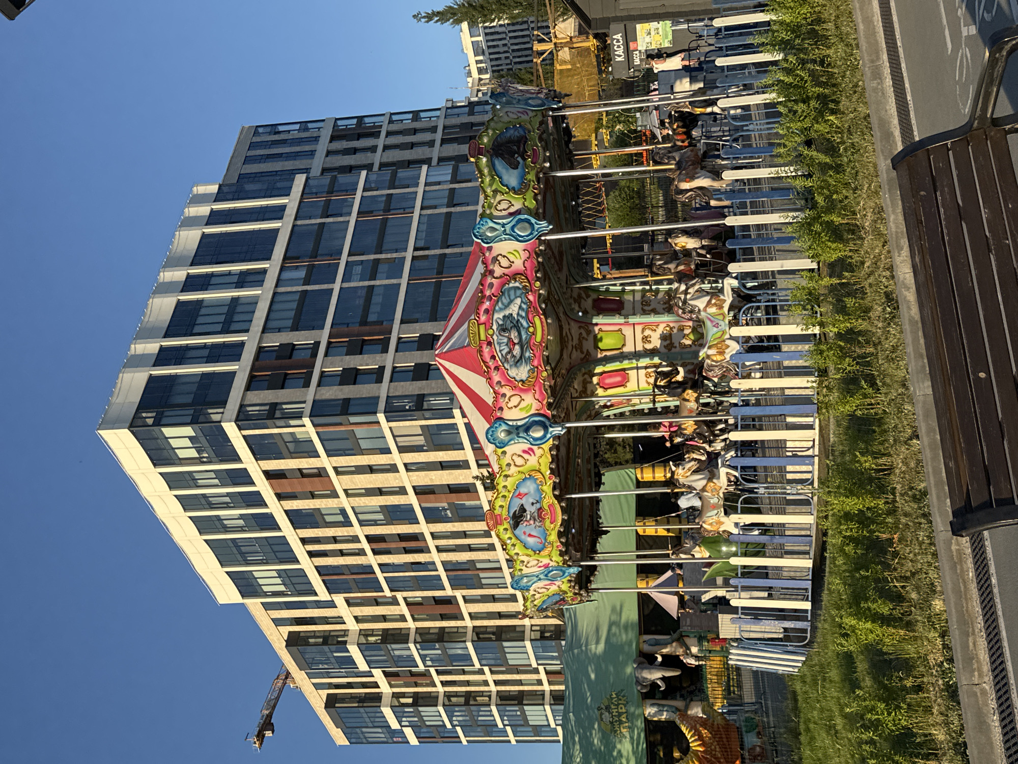 In Novosibirsk, Russia, a carousel spins in front of a modern, multi-story building with a facade of glass and beige stone. A construction crane is visible to the left of the building. The carousel is decorated with colorful panels featuring cartoon animals, including a cat and a horse. Children and adults are present, with some appearing to be riding the carousel. A green tent with elephants painted on it is set up behind the carousel. A sign with the word "КАССА" (Kassa, meaning "cash desk" or "ticket office" in Russian) is visible on a stand to the right of the carousel, indicating a ticketing or service area. The scene is set against a clear blue sky, with lush green grass and some bushes in the foreground, and a wooden bench in the lower right corner.