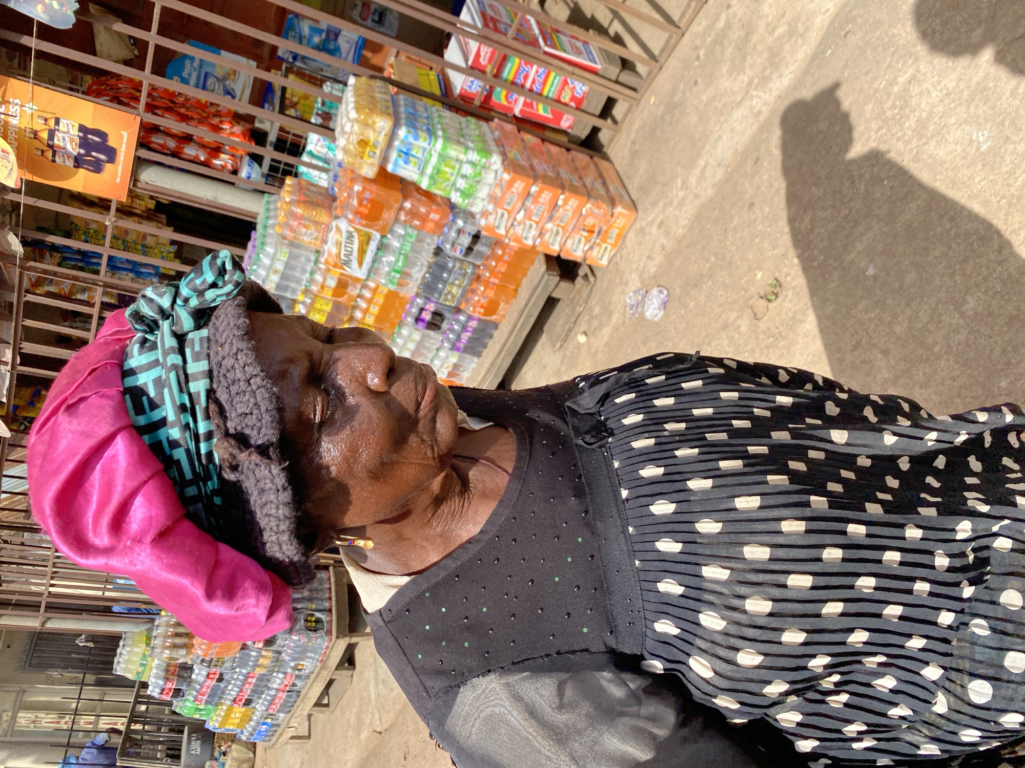 A medium shot captured outdoors in the city of Bukuru, Nigeria, shows an elderly Black woman standing in front of a store filled with beverages. The woman, wearing a bright pink headscarf over a knitted cap and a black dress with a polka dot pattern, is looking down and to her right. She has earrings and a necklace. Behind her, shelves are stocked with various brands of bottled drinks, including Maltina, Coca-Cola, and Pepsi, displayed on wooden pallets and metal shelving units. The scene is brightly lit by sunlight, casting shadows on the concrete ground.