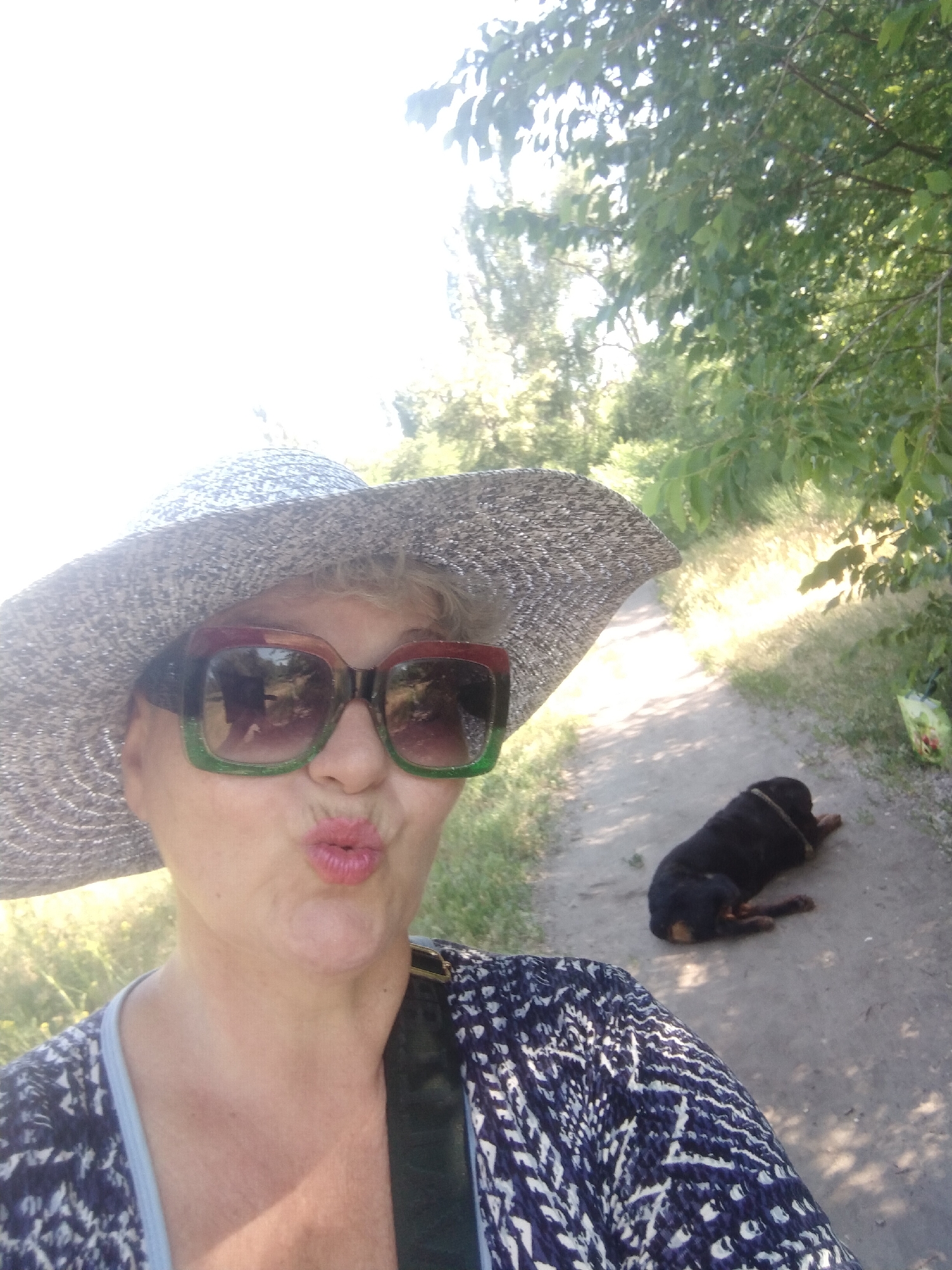 A woman wearing a wide-brimmed hat and sunglasses is taking a selfie outdoors on a dirt path. She is puckering her lips. A black dog lies on the path behind her. Trees and foliage are visible in the background. The scene is consistent with a park or natural area, potentially located in Horishni Plavni, Ukraine, given the context.