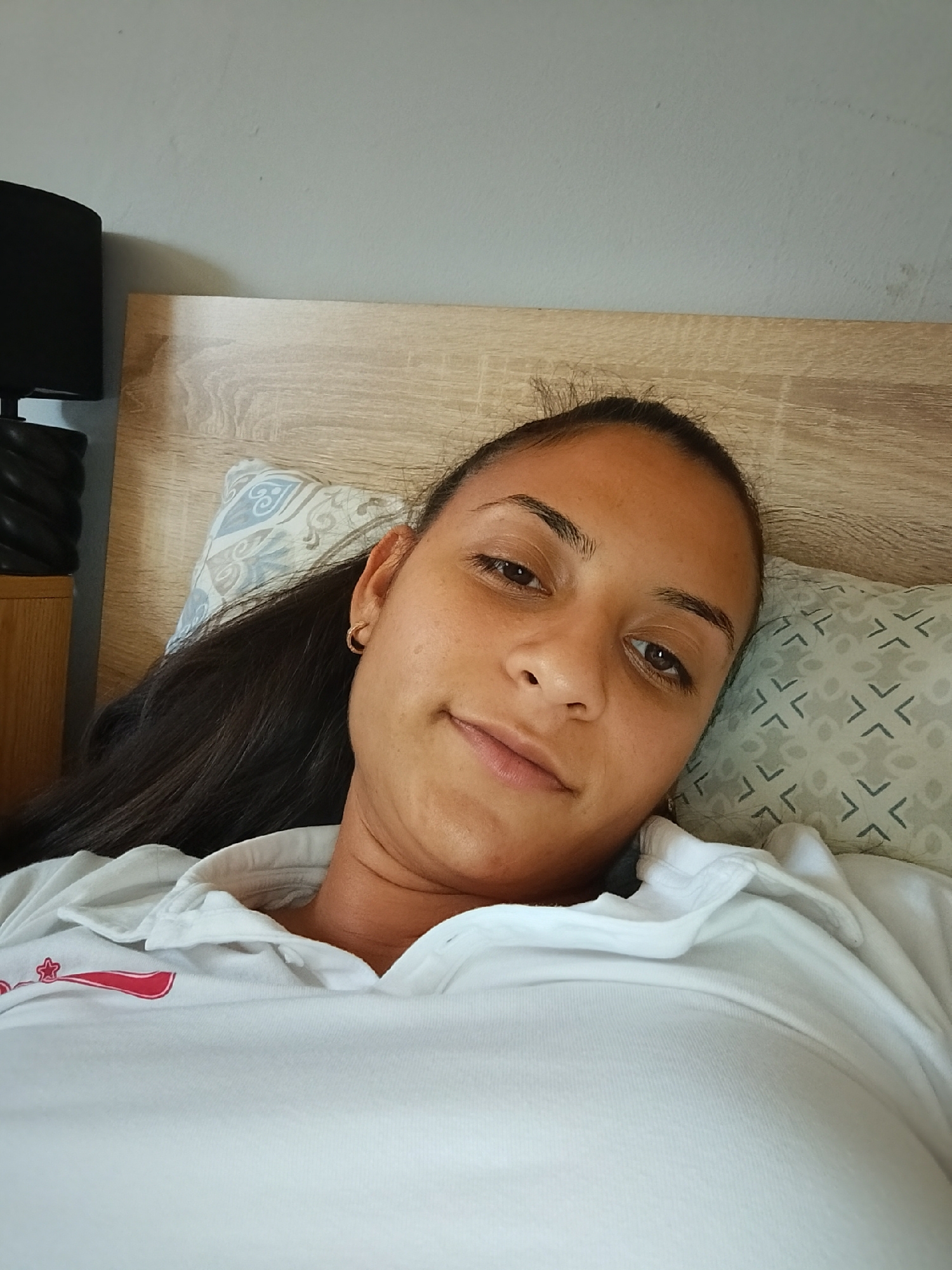A medium shot shows a woman with dark hair pulled back in a ponytail lying on her side in bed. She is wearing a white polo shirt. Her head rests on a pillow with a geometric pattern. Behind her, a wooden headboard is visible. To the left of the frame, part of a black table lamp with a spiral design is visible on a wooden nightstand. The background wall is a pale, neutral color. The scene appears to be a bedroom. The image does not contain any specific indicators of location within Yambol, Bulgaria.