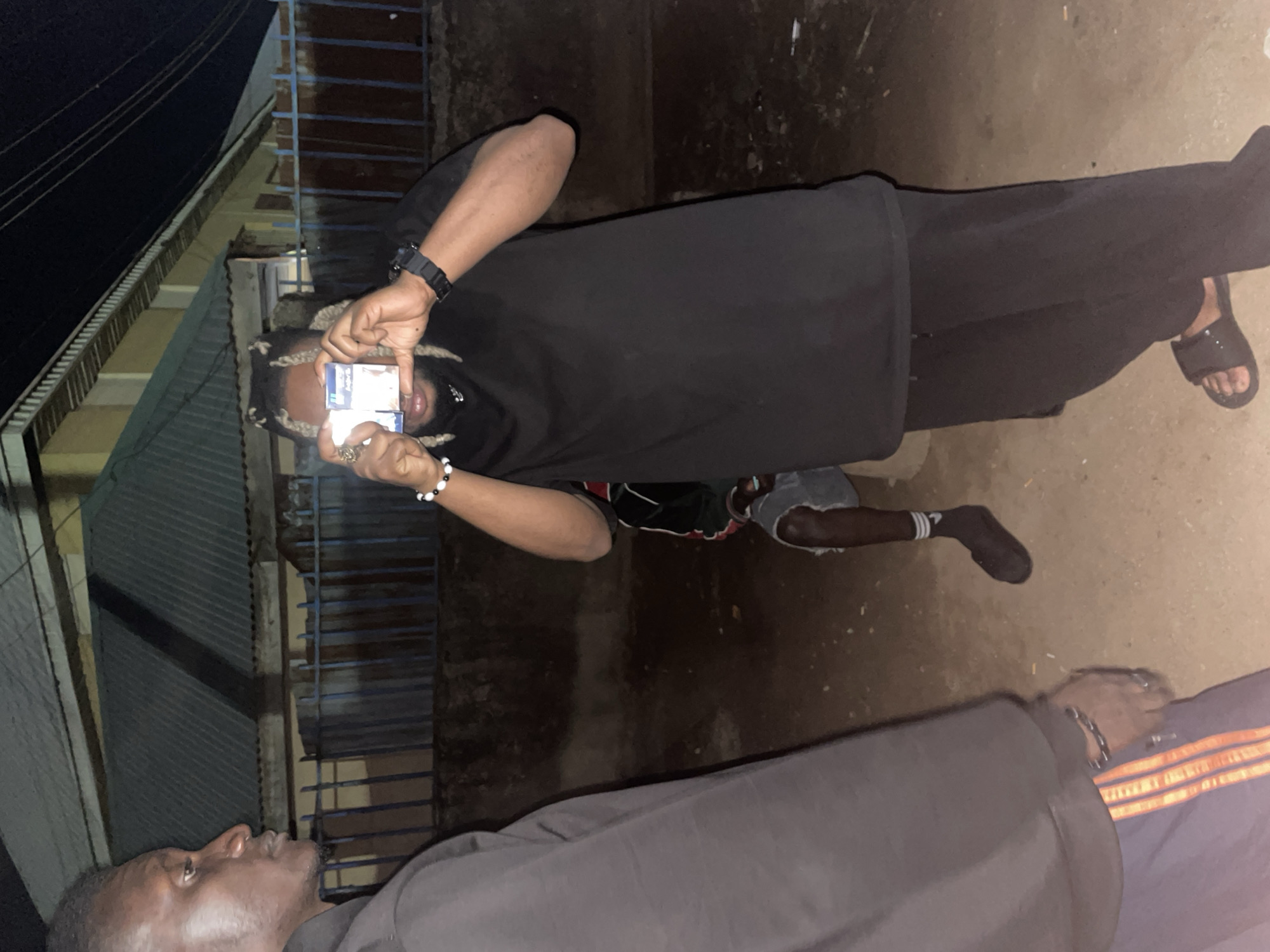 The image, taken at night in what appears to be an outdoor residential or street setting in Jos, Nigeria, shows three individuals in low light conditions. The primary subject, positioned centrally, is a person with dreadlocks and a beard, wearing a black t-shirt, black trousers, and black slide sandals on their feet. They have a black watch on their left wrist and a black and white beaded bracelet on their right. This individual is holding two small packs, likely cigarette boxes, up to their face, covering their mouth. Faint white text, possibly "Pall Mall" or a similar brand and "Cigarettes," is visible on the blue and white packaging of the top box.
To the bottom left, partially visible, is another person with dark skin, wearing a dark top, looking slightly upwards. In the background, between these two individuals, a third person, possibly a child, can be seen squatting or sitting, wearing shorts and dark socks.
The setting is characterized by corrugated metal fences or roofs in the background, suggesting a somewhat rustic urban or suburban environment. Overhead, some electrical wires are visible against the dark sky. The ground appears to be a mix of dirt and possibly some refuse. The overall atmosphere is casual, and the scene seems to capture an informal moment among the individuals.