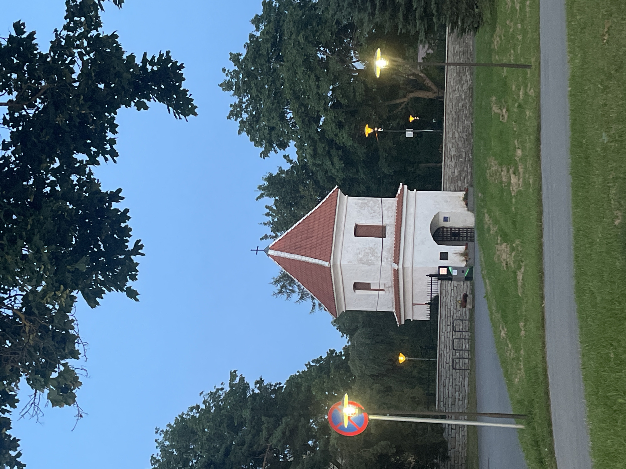 This image shows a small, white, two-story building with a red tile roof, topped with a cross, which is likely a historical tower or small church. The building is surrounded by a stone wall and lush green trees. In the foreground, there is a paved area with grass and a lamppost. A "no parking" sign is visible on the left side of the image. The sky is a clear blue, suggesting it is either early morning or late evening. The architectural style of the building, particularly the red tile roof and the white stucco walls, is characteristic of historic European architecture, and specifically, it resembles the Kiek in de Kök fortification tower in Tallinn, Estonia, which has a similar design and historical context.