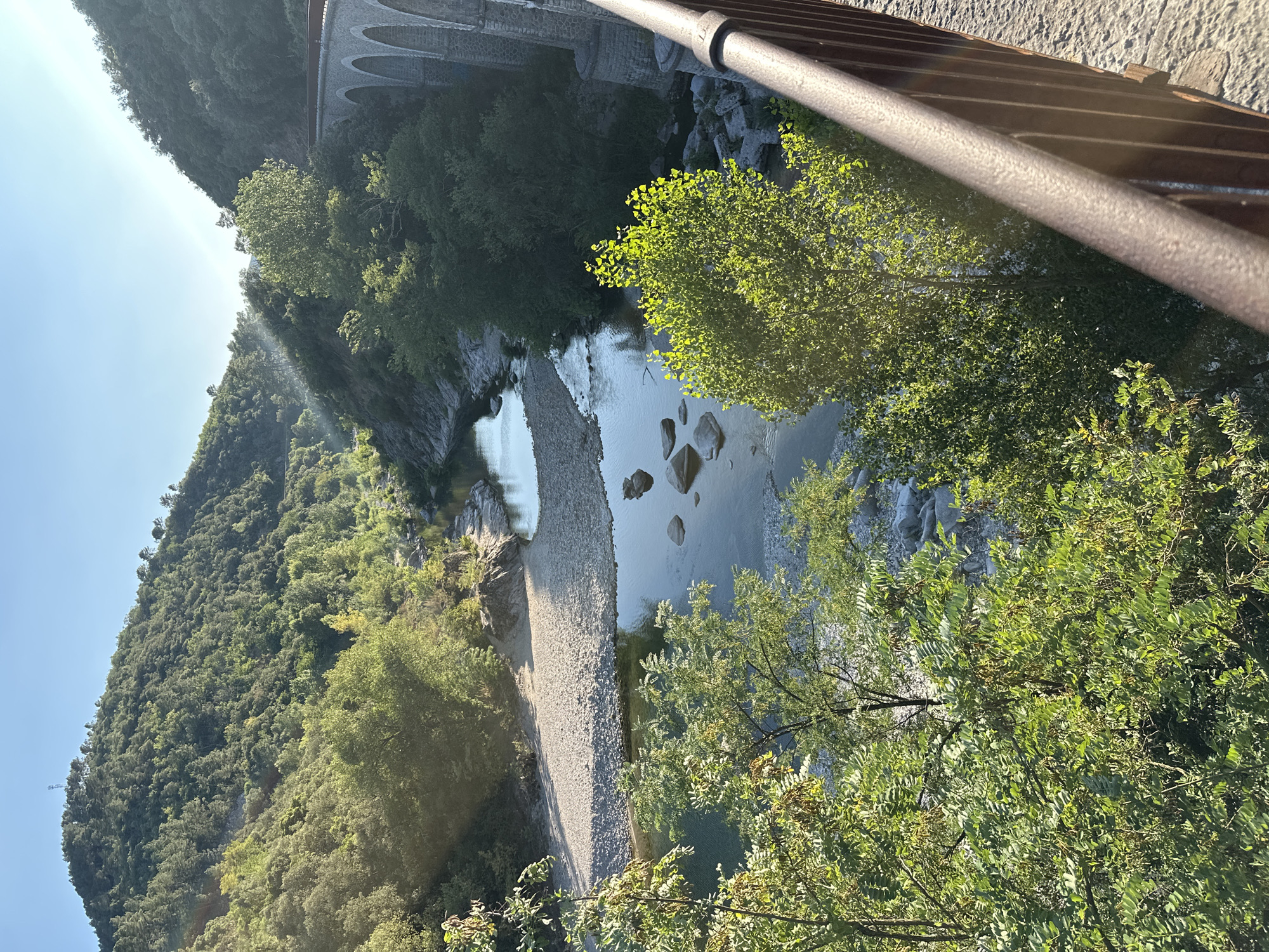 The image captures a scenic view of a river flowing through a valley, with a stone bridge arches visible on the right side of the frame. The river is surrounded by lush green trees and vegetation covering the steep hillsides. The sun is shining, casting a warm glow on the landscape, with some light rays piercing through the trees in the background. A gravelly riverbed is visible along the banks, with smooth stones scattered in and around the water. The bridge, constructed of stone, features multiple arches and appears to be an old structure, possibly part of a railway line given its design. The foreground is partially obstructed by a metal railing, suggesting the photo was taken from a vantage point, likely a bridge or a walkway. The overall impression is one of natural beauty and historical architecture harmoniously coexisting. The location is identified as Générargues, France, a commune in the Gard department.