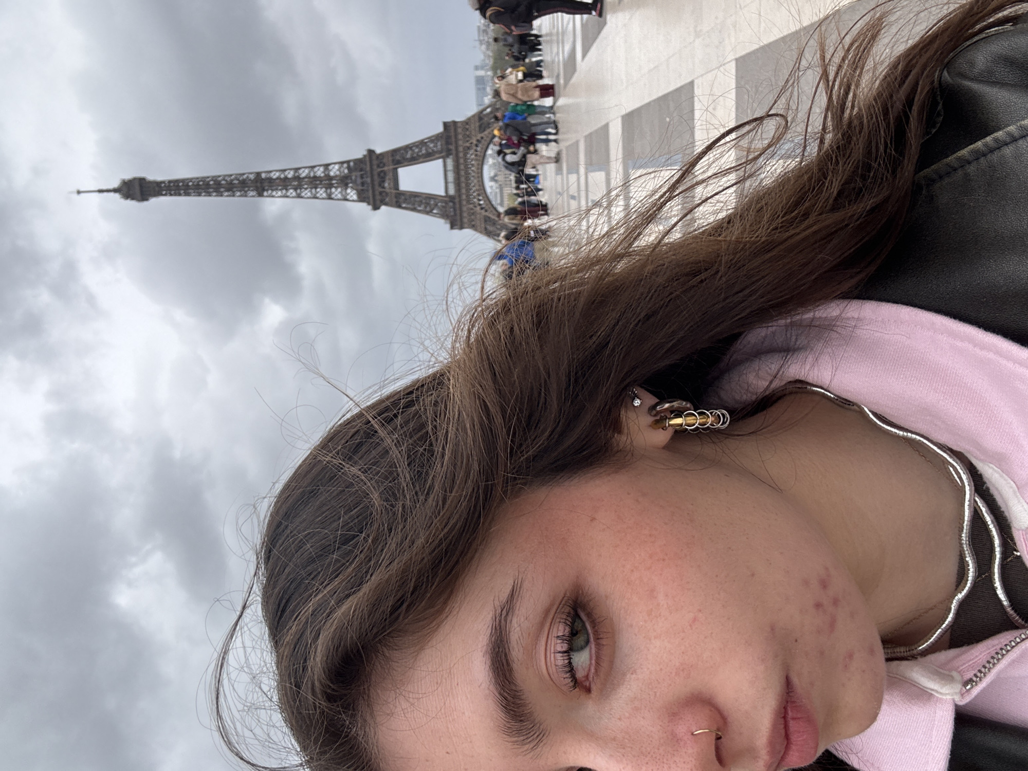 A photograph shows a young woman against the backdrop of the Eiffel Tower in Paris, France. The Eiffel Tower is in the background and off to the left; at its base are a crowd of people walking across the paved area. The sky is overcast. The woman has brown hair, a nose ring, and multiple earrings in her left ear. She wears a pink hooded sweatshirt and a black jacket. A silver necklace is visible.