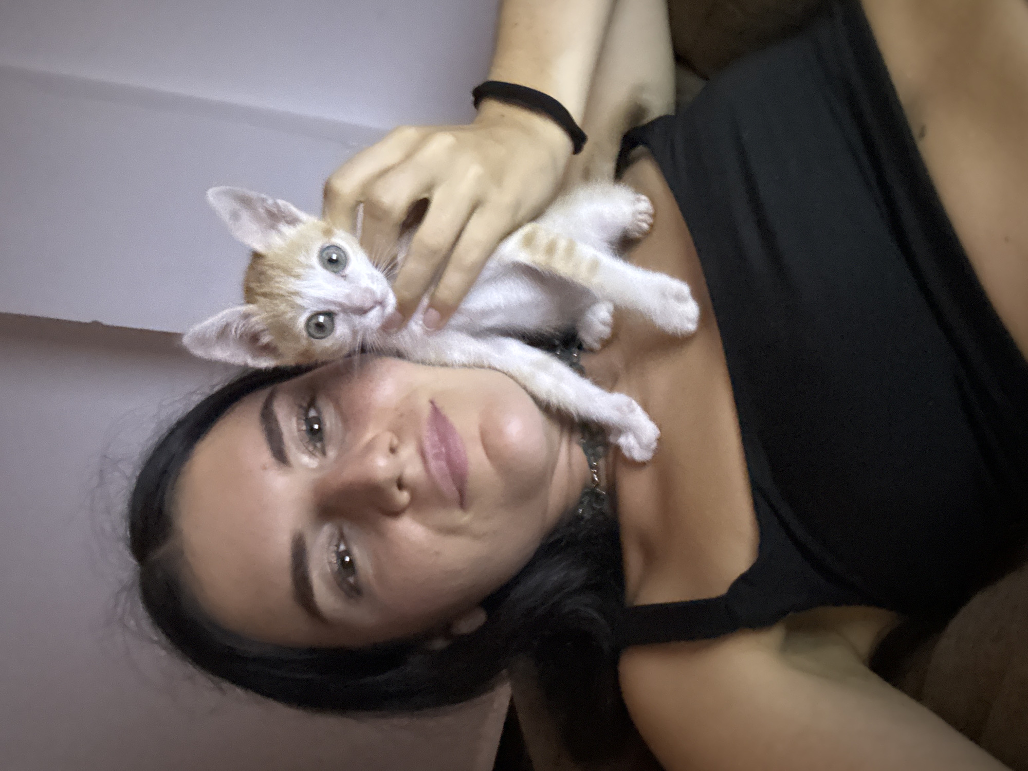A woman lies on what appears to be a brown or dark-colored upholstered surface, possibly a sofa or bed. She is oriented with her head towards the lower left corner of the frame and her body angled upwards towards the right. She has dark hair, fair skin, and is wearing a black sleeveless top. A small, light orange and white kitten is lying on her chest and neck. A hand, presumably belonging to the woman, is gently touching the kitten. The background is nondescript, with what appears to be a wall behind her that is a light shade. It is difficult to ascertain a specific location, particularly whether it is in Mũi Né, Vietnam, without more context. The scene appears to be indoors.