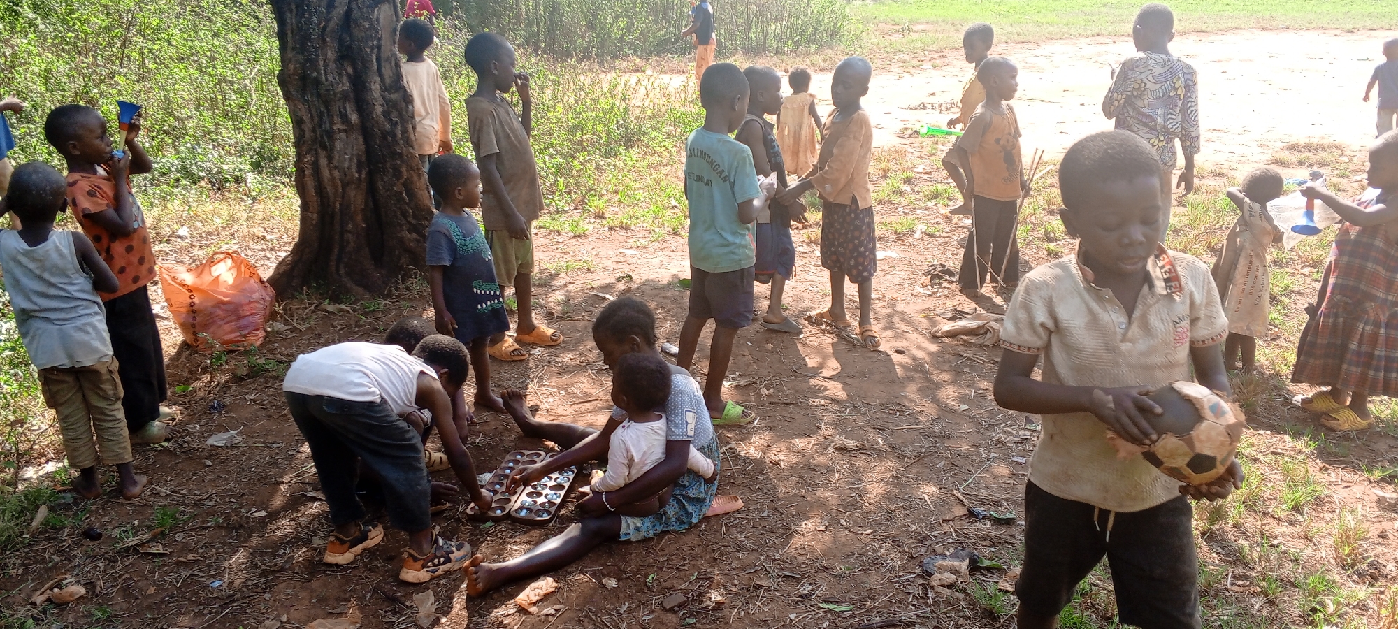 A sun-drenched, outdoor scene in Ngalula, Democratic Republic of the Congo, features a group of children, primarily of African descent, engaged in various activities. The environment consists of a dirt and grass ground, partially shaded by a large tree.
A boy in the foreground is holding a tattered soccer ball. To the left, several children gather around a game board on the ground, while another child sits, holding a toddler. A large orange plastic bag rests against the tree trunk. In the background, other children stand and observe, some holding cups or bottles. Their clothing is varied, ranging from shirts and shorts to dresses. The overall impression is one of simple, communal recreation.
