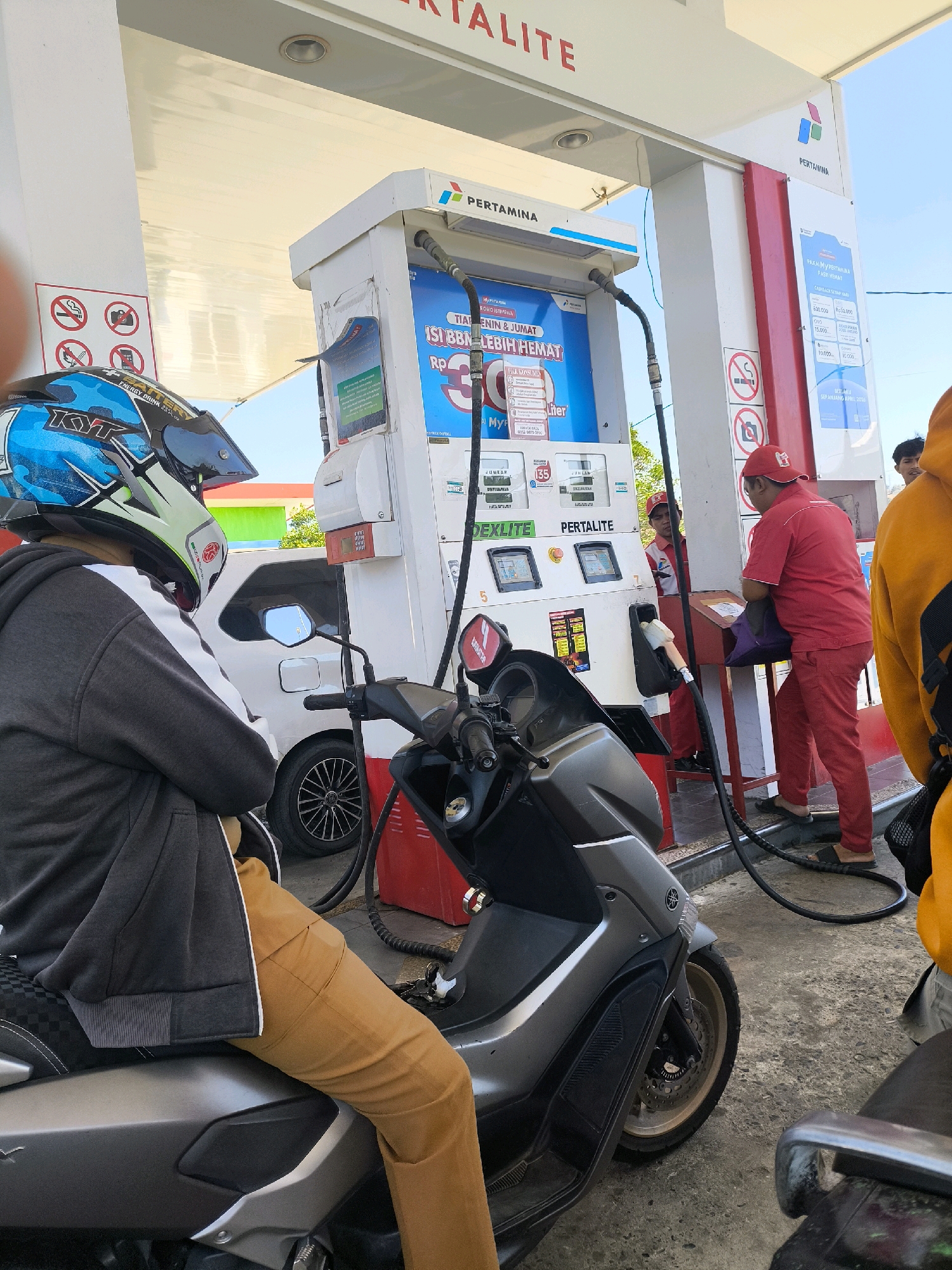 The image depicts a gas station in Kwandang, Indonesia, with a Pertamina gas pump prominently featured. A person wearing a black and gray hooded jacket and tan pants sits on a gray scooter, facing away from the camera and toward the pump. They are wearing a blue and white helmet with the "KYT" logo visible. Two gas station attendants are visible; one, wearing a red uniform and a red cap, is attending to a customer at the pump, while another, also in a red uniform, is in the background. A white car is parked behind the scooter. The environment appears to be an outdoor setting under a bright, clear sky. The image captures a typical scene at a gas station, with a focus on the refueling process.