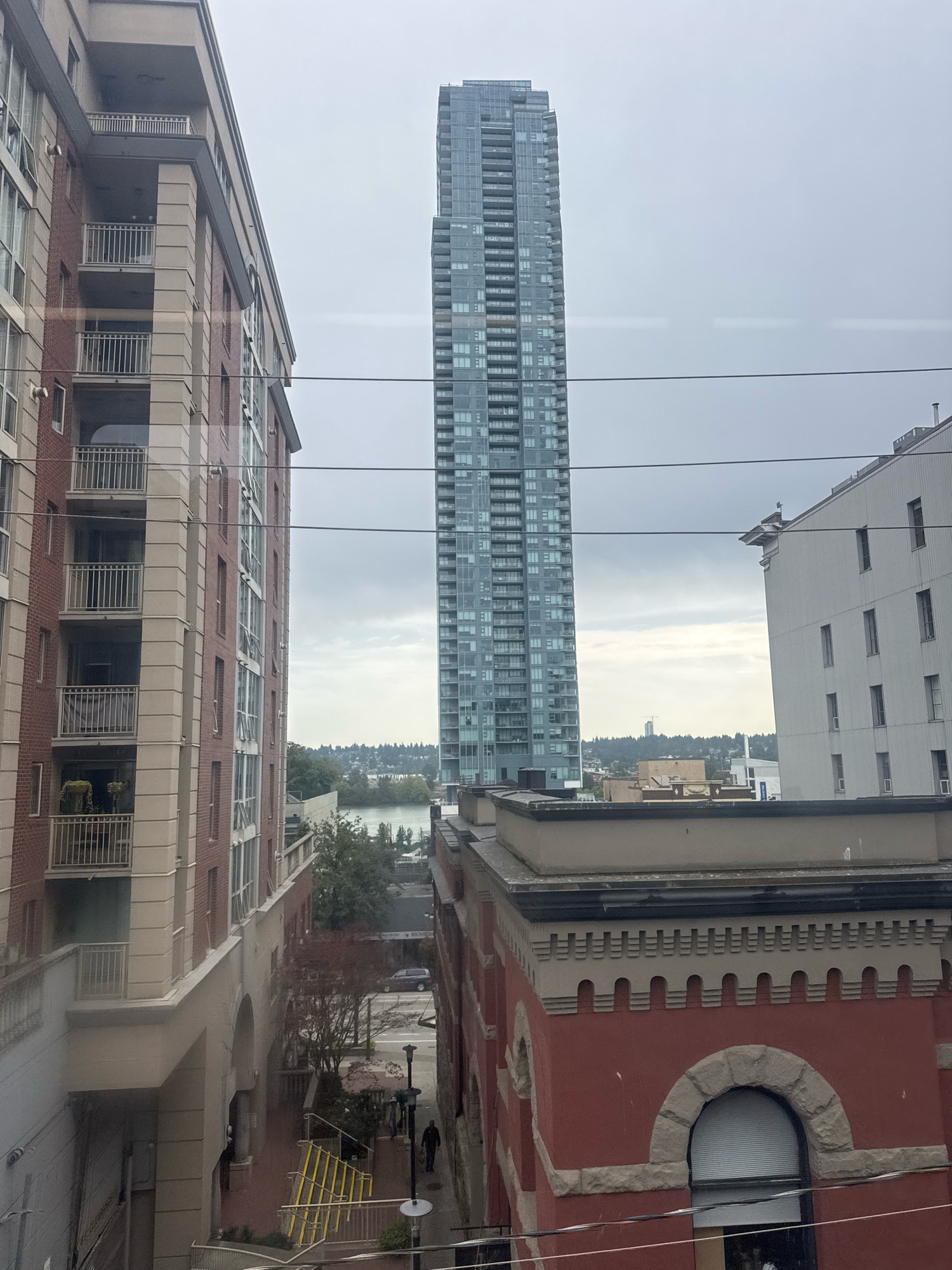 The image captures an urban scene in New Westminster, Canada, dominated by a cluster of buildings. On the left, a multi-story brick building with beige trim features balconies with metal railings. To its right, a very tall, slender skyscraper with a glass facade stands prominently. In the foreground and mid-ground, other buildings are visible, including a red brick structure with arched windows and decorative stonework, and a white building with multiple rectangular windows. In the distance, a body of water can be seen, bordered by a treed shoreline and a hazy skyline. Power lines traverse the sky across the frame. Two individuals are visible walking on a sidewalk or pathway between buildings in the mid-ground. The sky is overcast, suggesting a cloudy or diffused light condition.