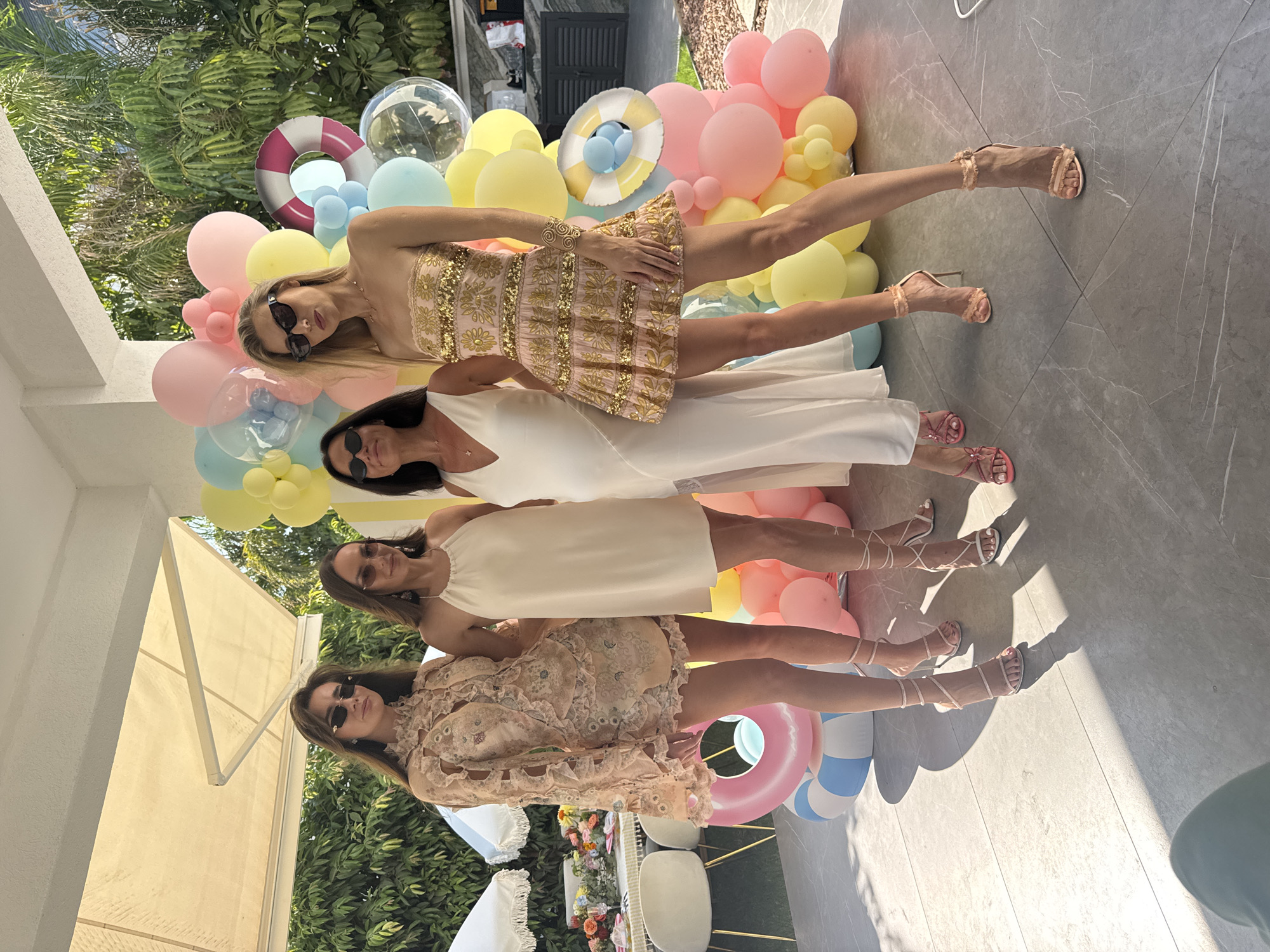 Four individuals stand in what appears to be an outdoor event space in Agios Tychona Community, Cyprus. The group consists of three adult females and one younger female. They are positioned in front of a large balloon arch in pastel colors, including pink, yellow, and blue, with a small inflatable life preserver and clear balloons. To the left of the group, a table is set with floral arrangements and umbrellas, suggesting a party or gathering. The ground is paved with large, gray tiles.
The individual on the far left, a younger female, wears a light peach-colored, ornate dress with intricate lace details and high neckline. She has long, dark hair and is wearing sunglasses and strappy heels.
The next individual to the right, an adult female, is dressed in a simple, sleeveless white dress and sunglasses, also with strappy heels.
The third individual, an adult female, stands in the middle, wearing a white V-neck dress. She has dark hair and is wearing sunglasses and strappy heels.
The individual on the far right, an adult female, is wearing a strapless, light pink dress adorned with gold embellishments and sequins. She has blonde hair, sunglasses, and strappy heels. She is also wearing a gold spiral bracelet on her right wrist.
The background includes lush green foliage, indicating a tropical or Mediterranean setting. The overall atmosphere suggests a celebratory occasion, possibly a bridal shower or birthday party, given the decorations and attire.