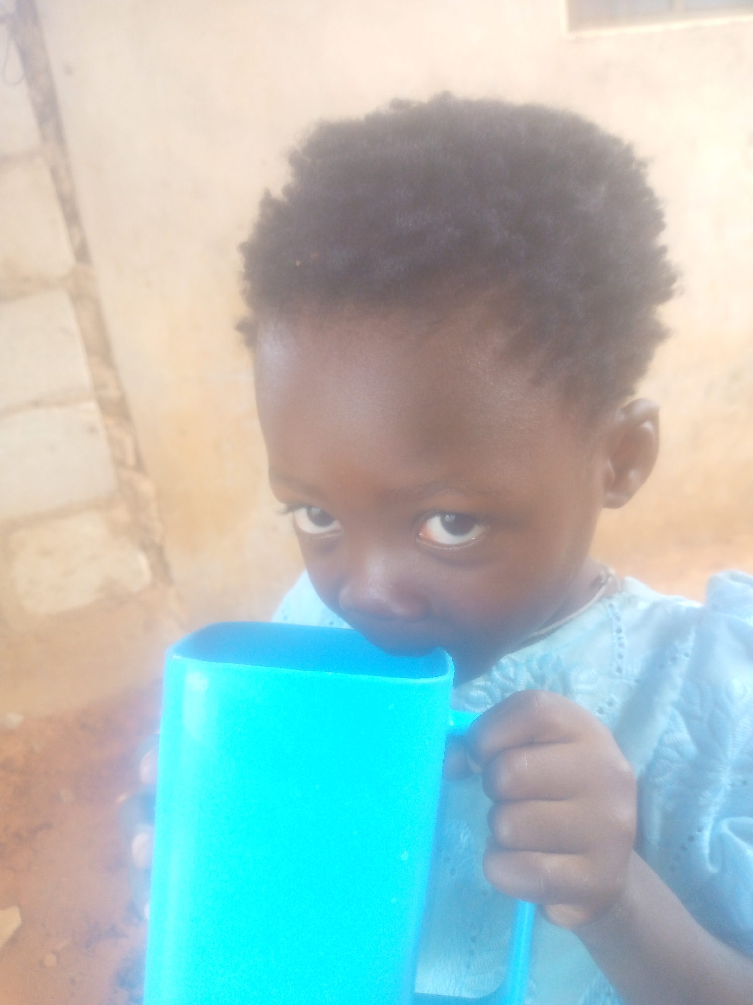 A young child with dark skin and afro-textured hair is drinking from a bright blue mug. The child is wearing a light blue, lace-patterned garment. The background is a blurred wall and ground. The location is not discernible from the image.