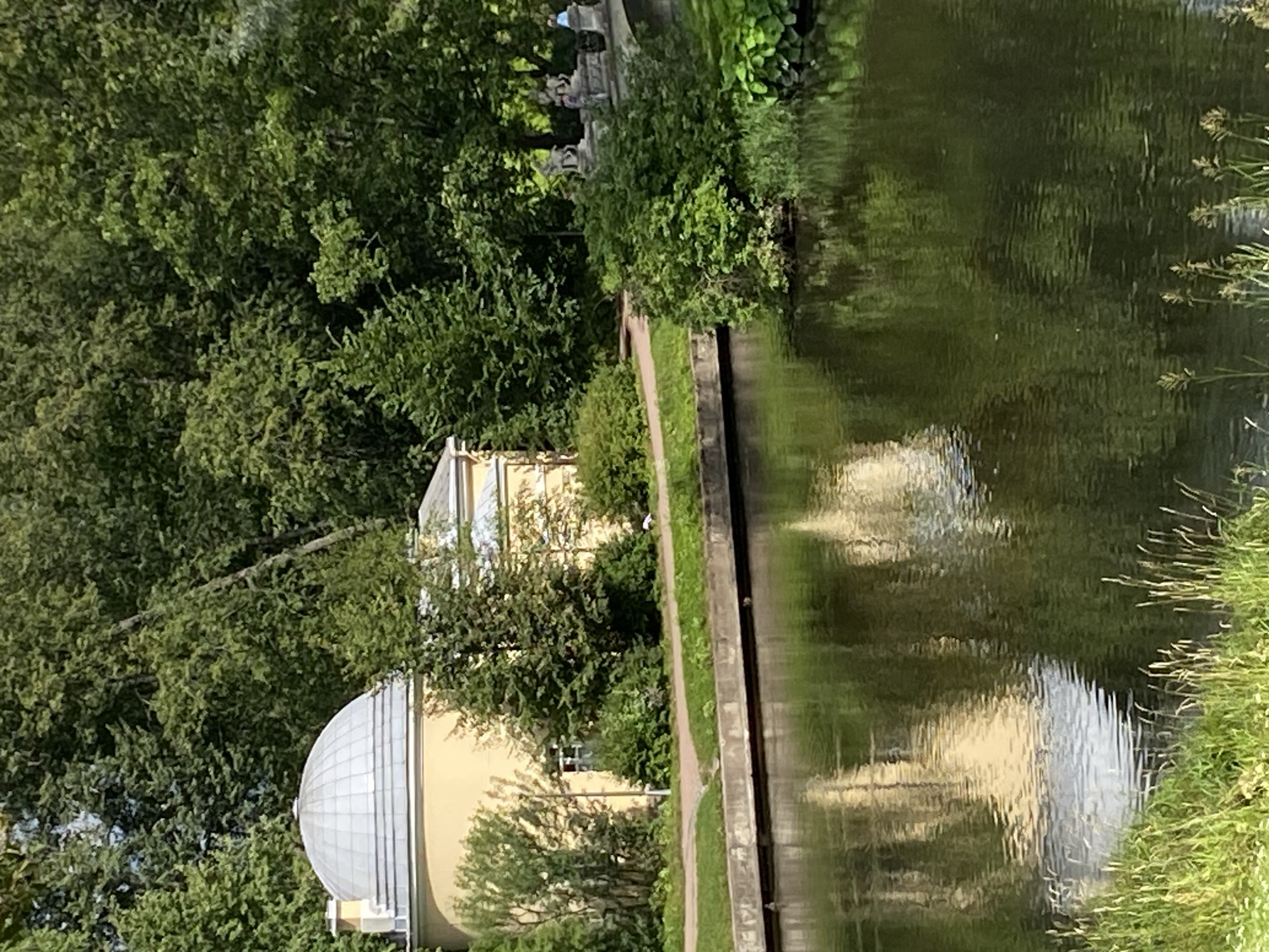 This is a photograph of a building with a domed roof, possibly a pavilion, situated by a body of water. The building is painted yellow and appears to be in a park-like setting with lush green trees and foliage. The water reflects the surrounding trees and the building. In the background, there is a bridge with sculptures on it. The overall scene suggests a serene and picturesque location. This scene depicts the Slav Library pavilion and the Agate Bridge in the Pavlovsk Park, located in Pavlovsk, Russia.
