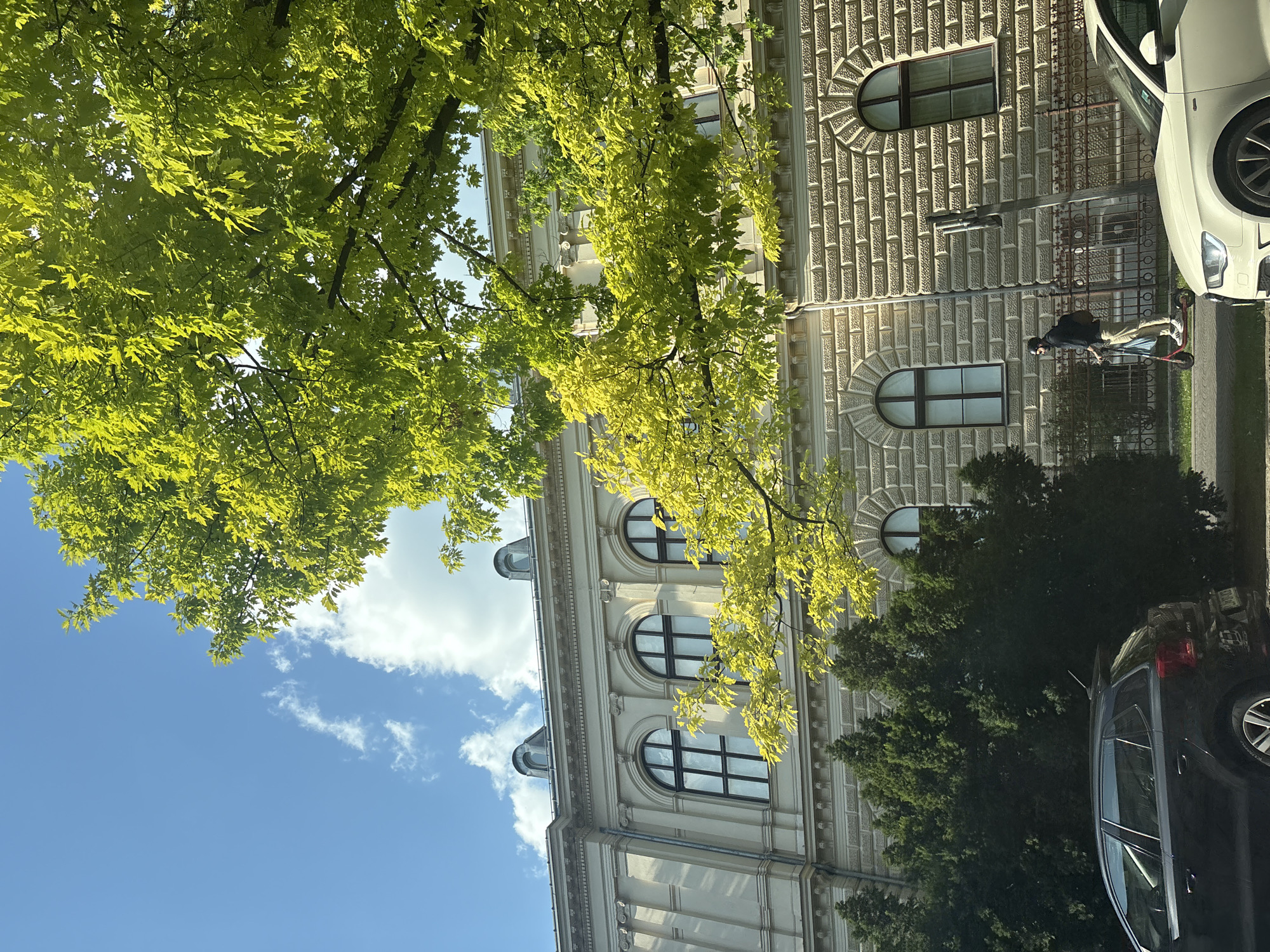 An eye-level, medium shot captures a street scene in a city, with a large, ornate building dominating the background. The building, constructed of light-colored stone with decorative elements and arched windows, features a mansard roof with dormer windows. In the foreground, a vibrant green tree with bright leaves partially obscures the building and the blue sky above, which is dotted with white clouds. To the left, a dark-colored car is parked, and to the right, a person is riding an electric scooter past a fence and more trees. A white car is partially visible in the lower right corner of the image. The lighting suggests it is daytime, with the sun casting shadows and highlighting the greenery.