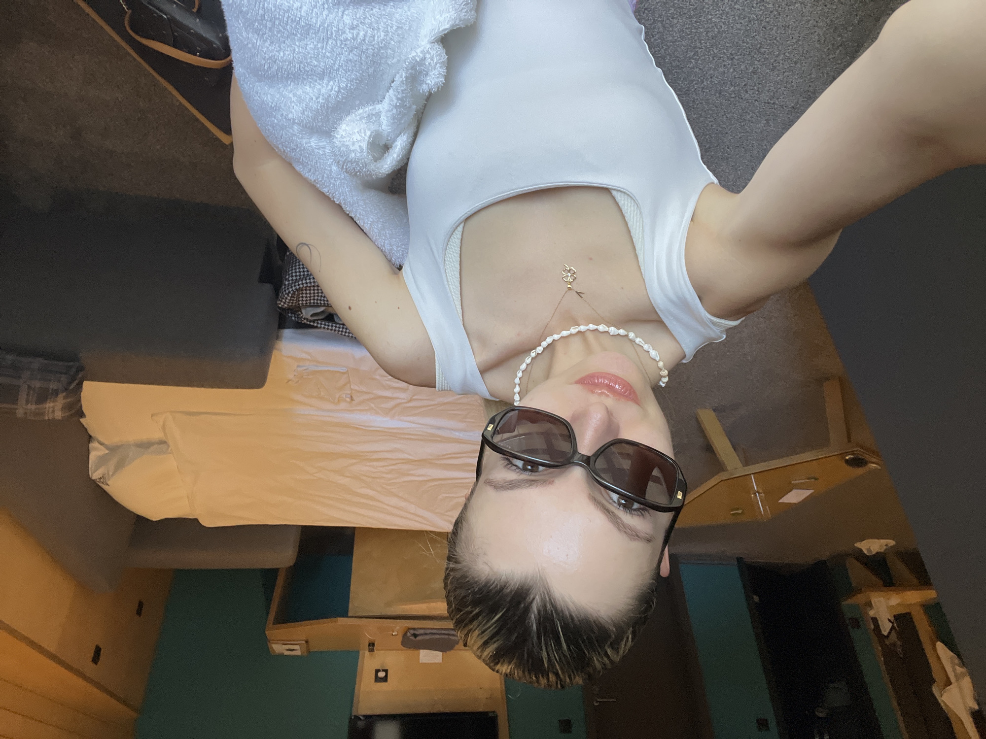 A woman is taking a selfie indoors. She wears dark sunglasses, a white top, and a pearl necklace. A towel is draped over her shoulder. A bed and furniture are visible in the background. The location is Unknown, Unknown.