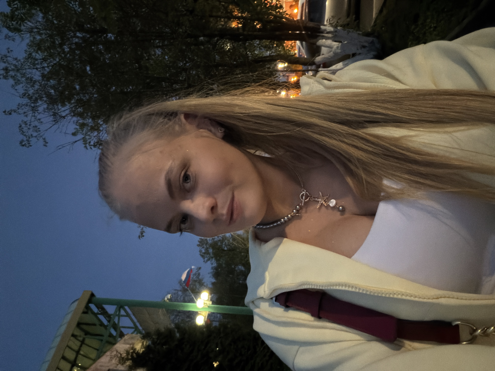 A young woman is pictured in a selfie. She has blonde hair and is wearing a white tank top, a light yellow zippered sweatshirt, and a silver necklace with a starfish charm. A maroon strap is visible on her left shoulder.
In the background, the Russian flag is displayed on a flagpole near a pedestrian bridge or walkway. The location appears to be a park or urban area in Izhevsk, Russia, based on the architecture and the presence of the Russian flag. There are trees and lights in the background, suggesting it is dusk or evening.