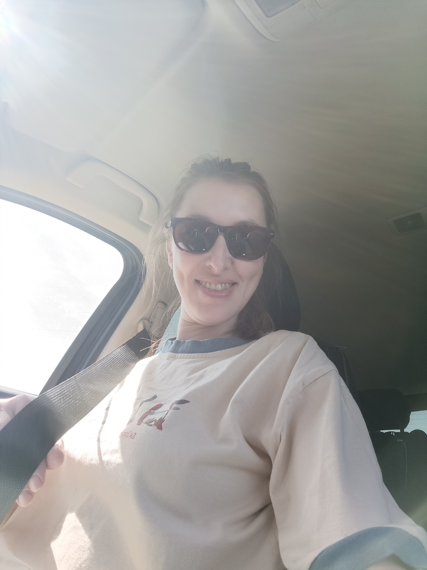 A woman is photographed from a low angle inside a car. She is wearing sunglasses, a tan t-shirt with a blue collar and cuffs, and a seatbelt. The background shows the interior of a car with the window, headrest, and ceiling visible. The location cannot be determined to be in Chaikovsky, Russia, from the provided image.