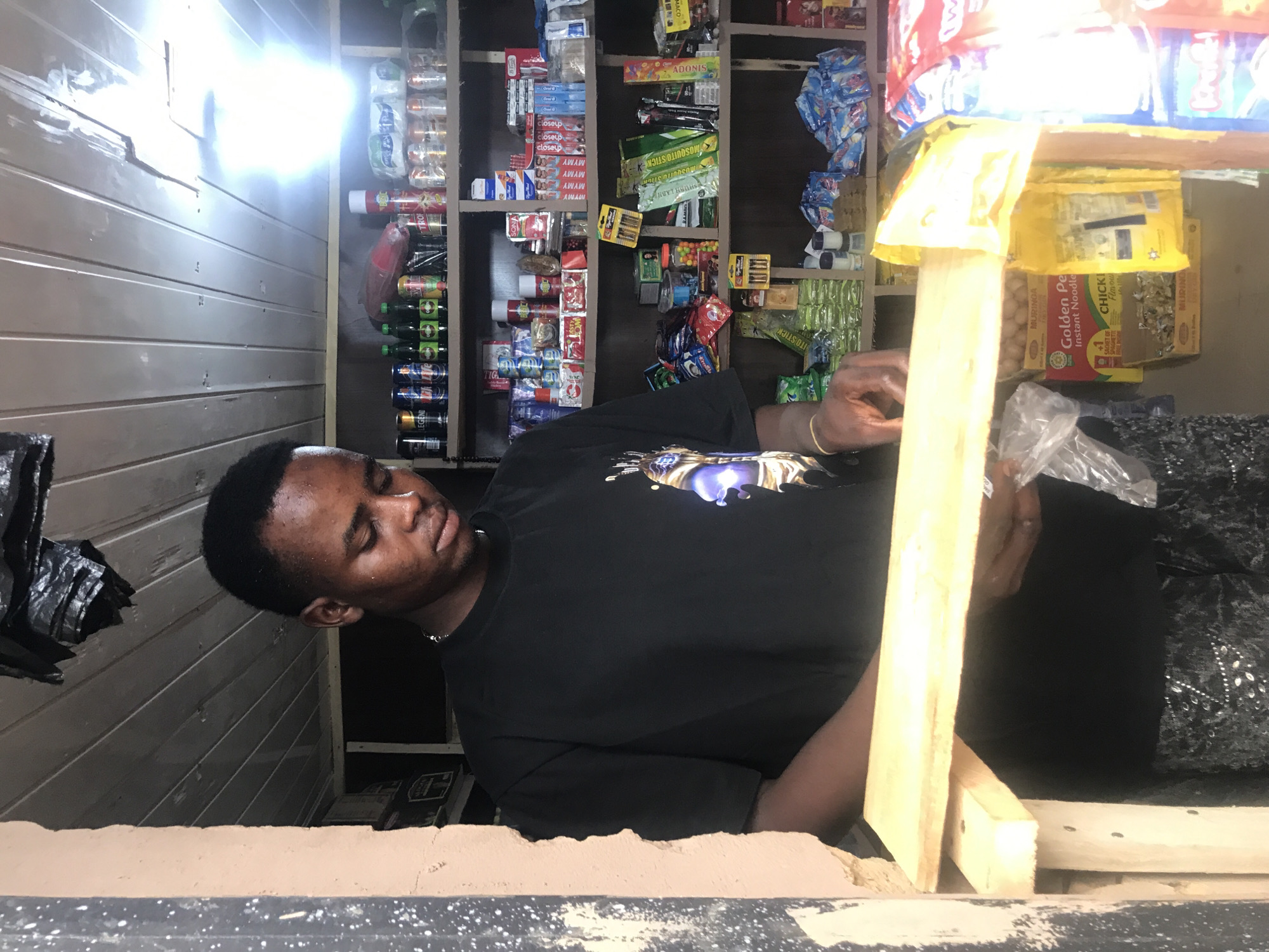 In New Stadium, Nigeria, a young Black male stands behind a wooden counter in a small convenience store. He is wearing a black t-shirt with a graphic design on the front. The shelves behind him are stocked with various products, including canned drinks, toiletries, snacks, and other household items. The store appears to be dimly lit with overhead fluorescent lighting. The man is looking down and appears to be engaged in an activity related to the counter or the items on it, with his hands positioned as if handling or arranging products. The environment suggests a local retail setting with a variety of goods for sale.