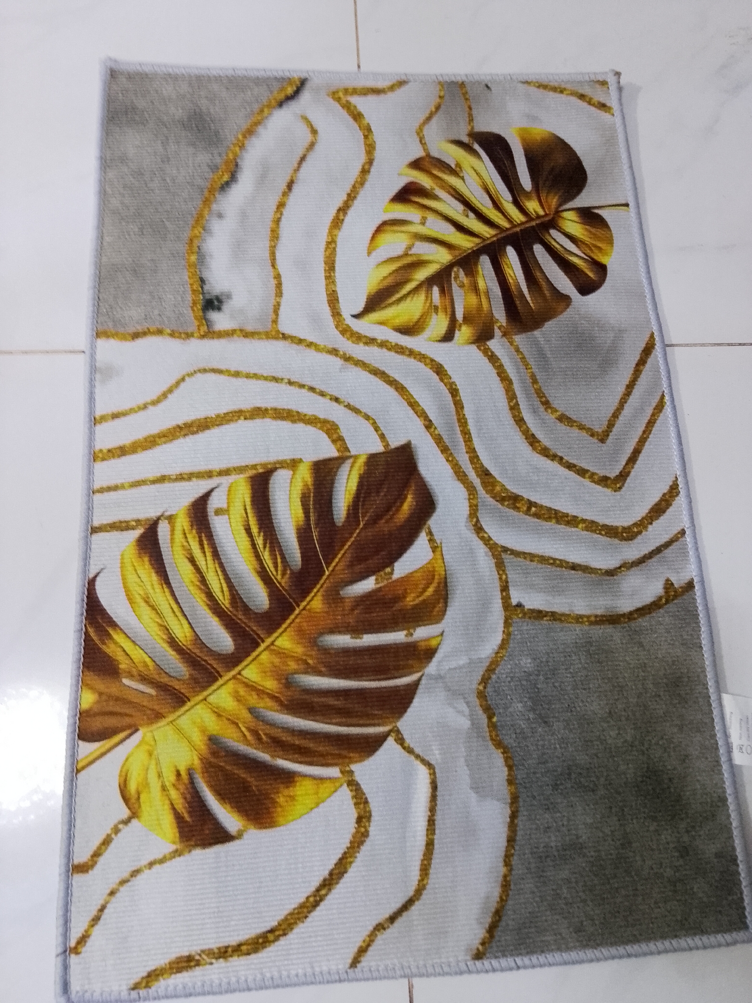A rectangular mat or rug is depicted, laid on a white tiled floor. The mat features a prominent design of two large, golden Monstera leaves, which appear to have a metallic or shimmering texture. These stylized leaves are placed on a background that combines patches of light gray with off-white areas. Wavy, shimmering golden lines traverse the mat, abstractly outlining shapes and complementing the leaf motifs. The mat has a neat, light gray stitched border along its edges.
There are no people or specific activities taking place; the image is a static shot of the decorative mat. The setting appears to be indoors, likely in a home or commercial space in Davao City, Philippines, given the location context. The light source is overhead, creating soft shadows and highlighting the golden elements of the design.
In the bottom right corner of the mat, a small white label or tag is partially visible. The text "OEKO-TEX" is clearly readable, indicating a certification for textile products. Below this, small, indistinct care symbols (such as washing or drying instructions) can be seen. The overall aesthetic of the mat is modern and elegant, characterized by its golden and grey color palette.