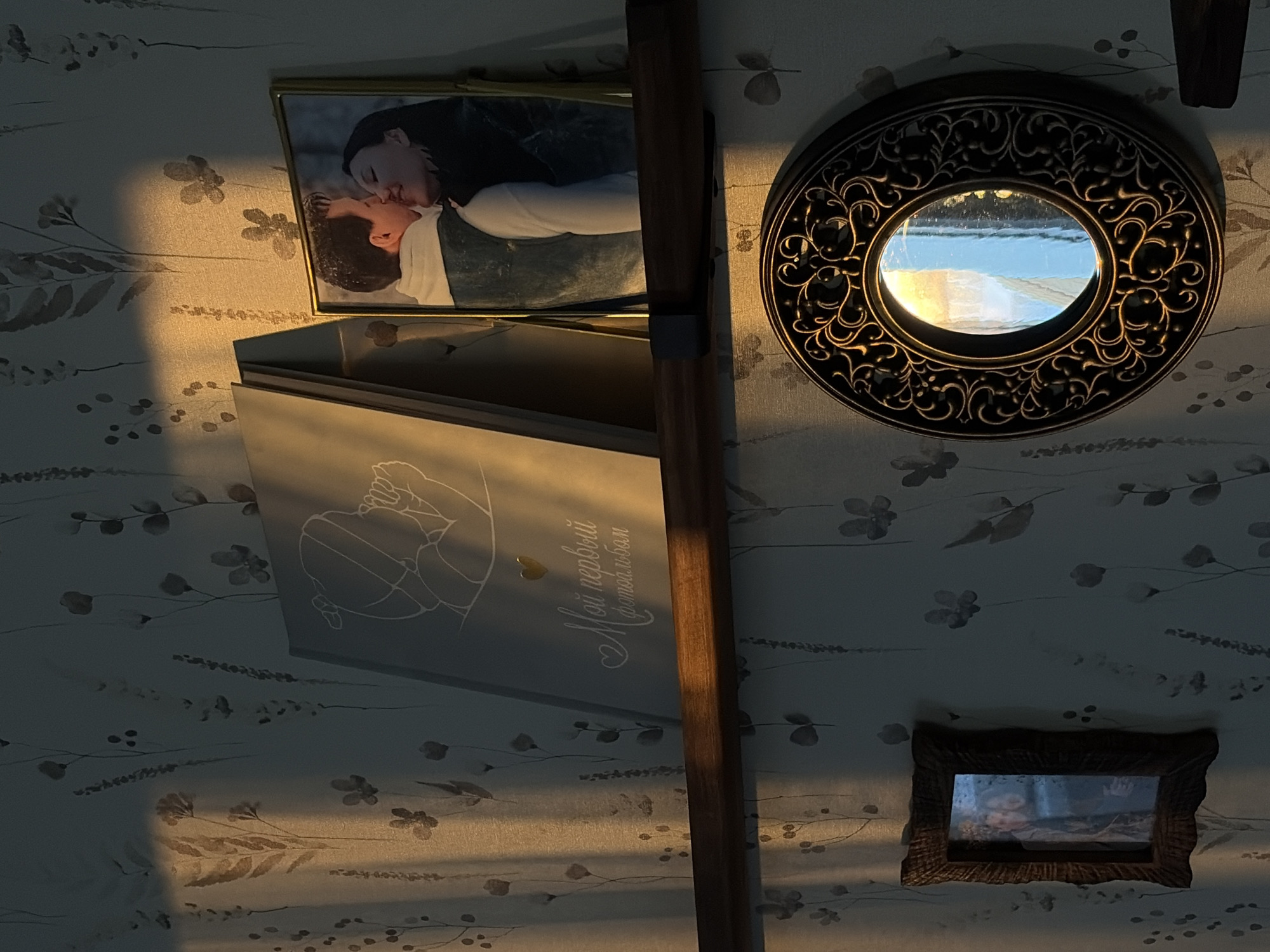 The image depicts an interior scene, likely a private residence in Chaikovsky, Russia, characterized by specific decorative elements. A patterned wallpaper, featuring muted beige and grey botanical motifs on a light background, covers the wall. Strong, warm light, presumably sunlight, enters from the left, casting elongated shadows and illuminating the upper left portion of the wall and objects.
A dark wooden shelf with metal brackets is mounted across the middle of the frame. On this shelf, from left to right, are:
1. A light grey book or photo album titled "Мой первый фотоальбом" (My first photo album) in Cyrillic script, with a small golden heart graphic and a minimalist line drawing of a baby. It rests leaning against the wall.
2. A gold-framed photograph featuring a man and a woman in a close-up, kissing. The man, with short dark hair, wears a white hooded garment and a dark outer layer. The woman has long dark hair and dark clothing. The background suggests an outdoor, possibly snowy, environment.
Below the shelf, affixed to the wallpaper, are:
1. On the left, a small, dark wooden picture frame with an irregular, rustic edge. The framed image is indistinct but appears to contain two figures.
2. On the right, a round mirror with a highly ornate, dark metallic frame featuring intricate scrollwork patterns. The mirror's reflection shows a bright, blue sky and what appears to be a window frame or an external light source.
