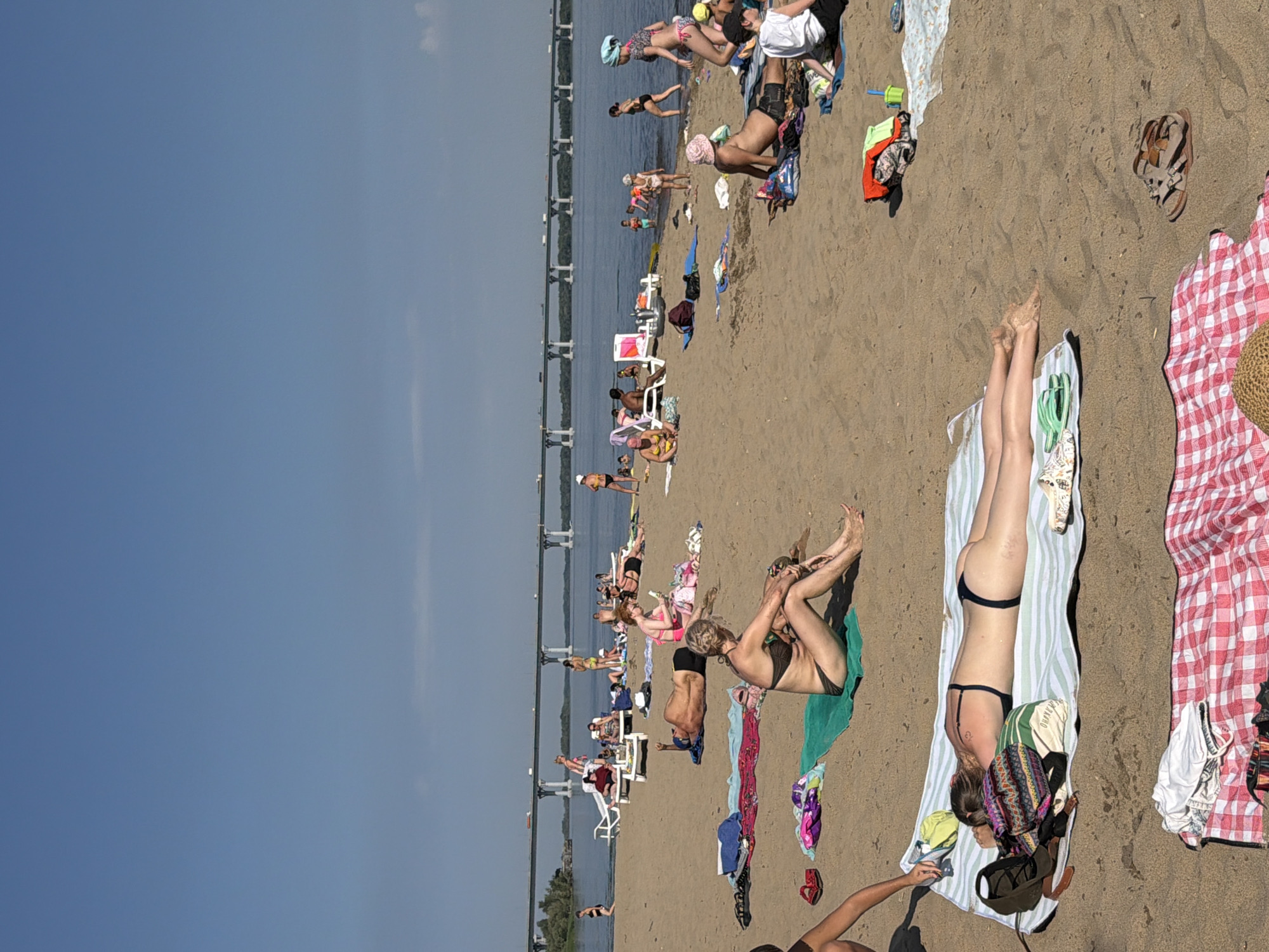 A wide-angle, eye-level shot captures a busy beach scene on a sunny day. The sandy shore is populated with numerous people in swimwear, engaged in various beach activities. In the foreground, a person lies supine on a striped towel, while another individual, also in a bikini, sits on a green towel to the right. Further back, more beachgoers lounge on towels and chairs, with some people wading or playing in the shallow water. In the background, a large bridge spans the wide river, with vehicles visible crossing it. The sky is clear and blue, indicating fair weather. The overall scene depicts a lively summer day at a riverside beach, likely within the Volzhskiye Dali area of Russia, characterized by recreational activities and a relaxed atmosphere.