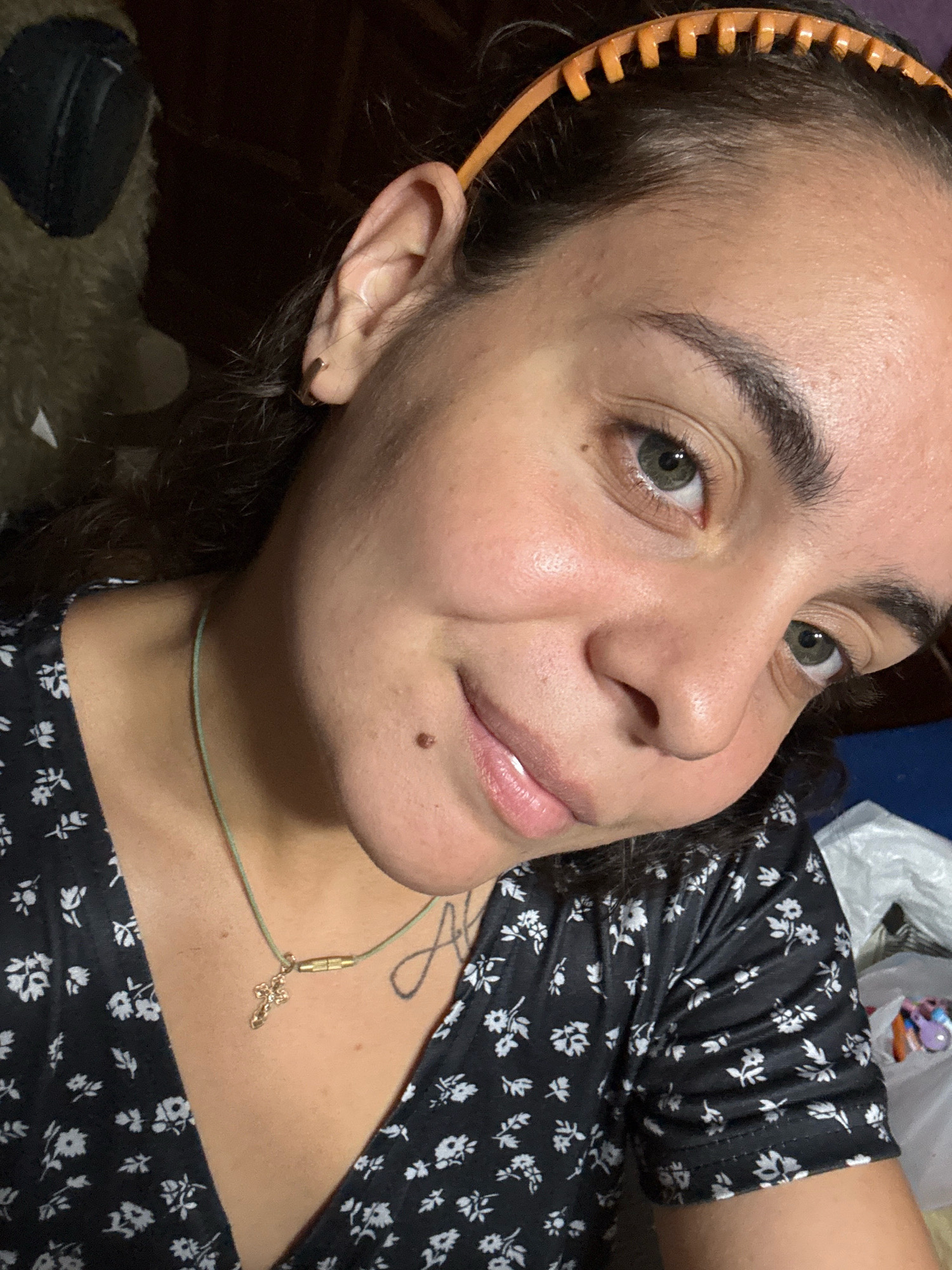 The image is a close-up selfie of a young woman with dark hair, wearing a black floral print top and a thin green necklace with a gold cross pendant. She has a mole on her right cheek and is wearing a light-colored headband with orange teeth. Her eyes are a greenish-gray color. The background is blurred, showing what appears to be a dark, possibly wooden surface to the left, and a blue surface and white plastic bag to the right. There is no discernible information in the image that indicates a specific location in Novocheboksarsk, Russia.
