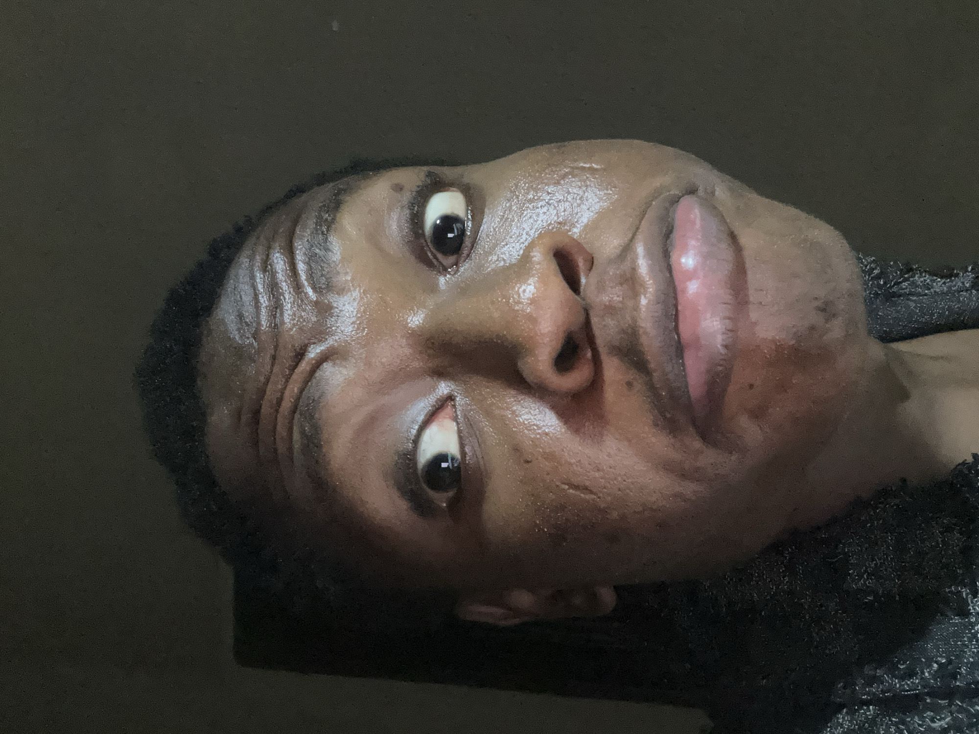 A close-up, low-angle shot depicts a man with dark skin. The individual appears to be sweating, exhibiting a slightly bulging eye expression. The background is obscured and dark, revealing only a section of black fabric on the right side.