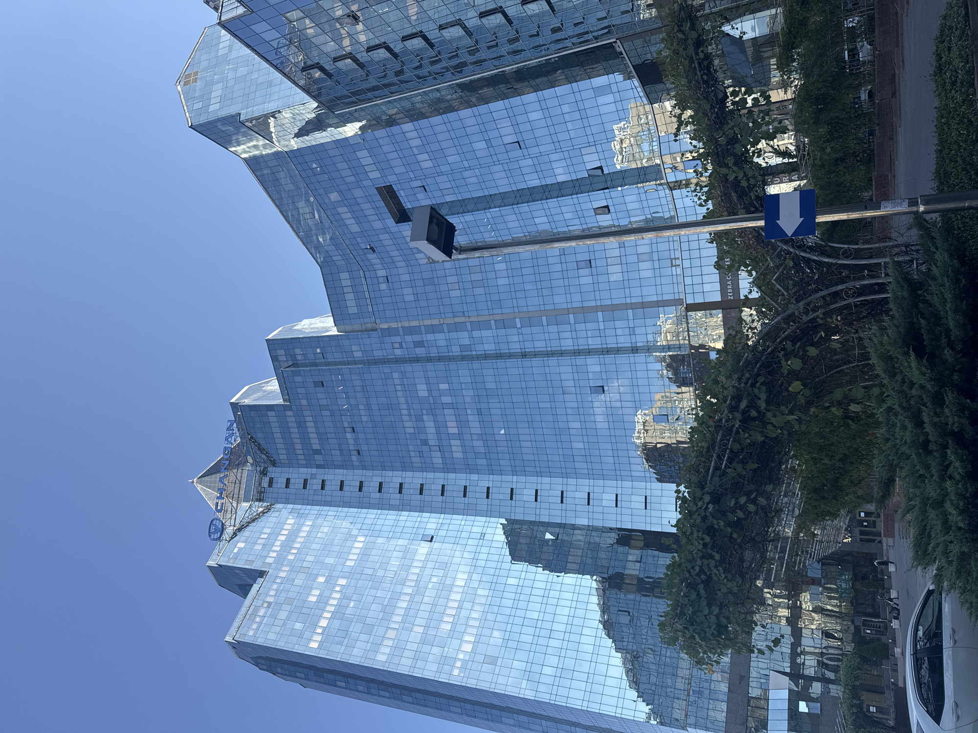 A low-angle, medium shot captures the modern glass facade of the Changan Sky Tower skyscraper in Almaty, Kazakhstan. The building's reflective blue glass surfaces mirror the clear, bright blue sky and surrounding structures. The skyscraper's angular design features multiple staggered sections, topped with the blue "CHANGAN" logo and a pointed apex. In the foreground, lush green bushes and an arched metal structure with trailing vines partially obscure the lower portion of the building. A street lamp with a directional sign pointing left is visible in the middle ground. A white sedan is parked on the left side of the image, and a few pedestrians are seen in the distance near the building's entrance. The overall impression is one of urban modernity and architectural dynamism under a sunny sky.