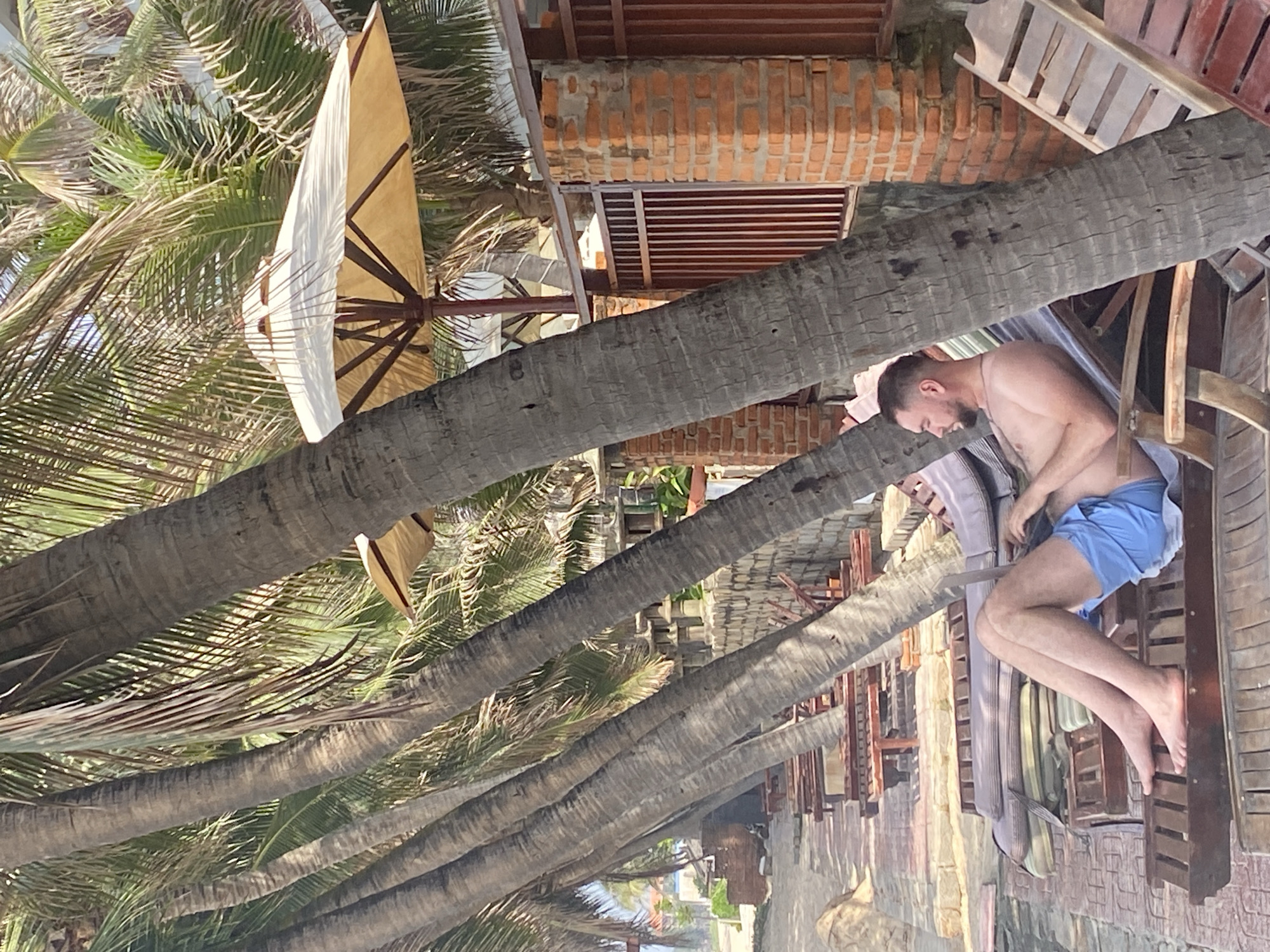 In Phu Thuy Ward, Vietnam, a man is lying on a lounge chair under the shade of palm trees on a beach. He is wearing blue shorts and appears to be using a laptop. The lounge chair is made of wood and has cushions. In the background, there are more lounge chairs, a brick wall, and a fence. The weather appears to be sunny.