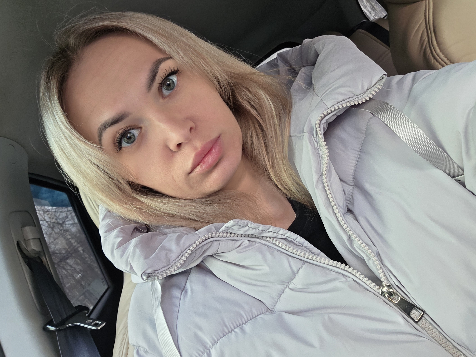 The image depicts a forensic, close-up view of a single subject, an adult female, situated inside what appears to be a passenger vehicle in Chaikovsky, Russia. The female, possessing long blonde hair and light-colored eyes, directly faces the camera with a neutral expression. She is wearing a light grey or silver puffer jacket, partially zipped, over a black undergarment. A car's interior is evident, featuring a beige or light brown seat to her right, and a seatbelt mechanism visible on the upper left. Through a vehicle window in the upper left background, blurred bare tree branches are discernible, suggesting an outdoor urban or suburban environment during a colder season. No other individuals or significant actions are present within the frame.
