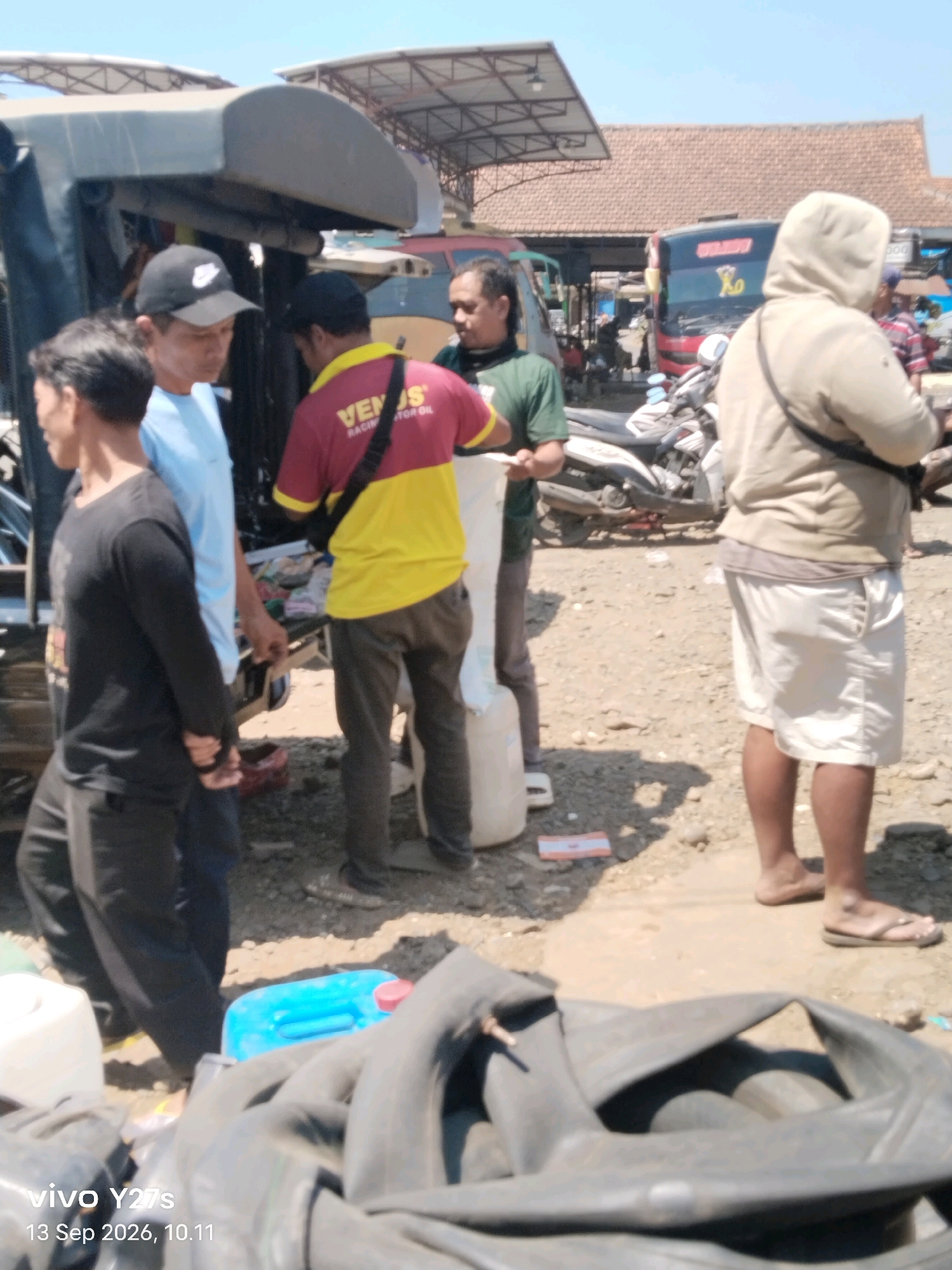 In Wonosido, Indonesia, on September 13, 2026, at 10:11 AM, a group of individuals is gathered outdoors. The scene appears to be a market or roadside area, with a bus visible in the background and several motorcycles parked. In the foreground, a person in a black long-sleeved shirt and pants stands next to another person wearing a light blue t-shirt. A third person in a red and yellow shirt is interacting with them. Further to the right, a person wearing a light brown hooded sweatshirt and white shorts is present. There are various objects scattered around, including large plastic containers, one of which is blue, and several tire tubes. The ground is dirt and gravel. The lighting suggests a sunny day.