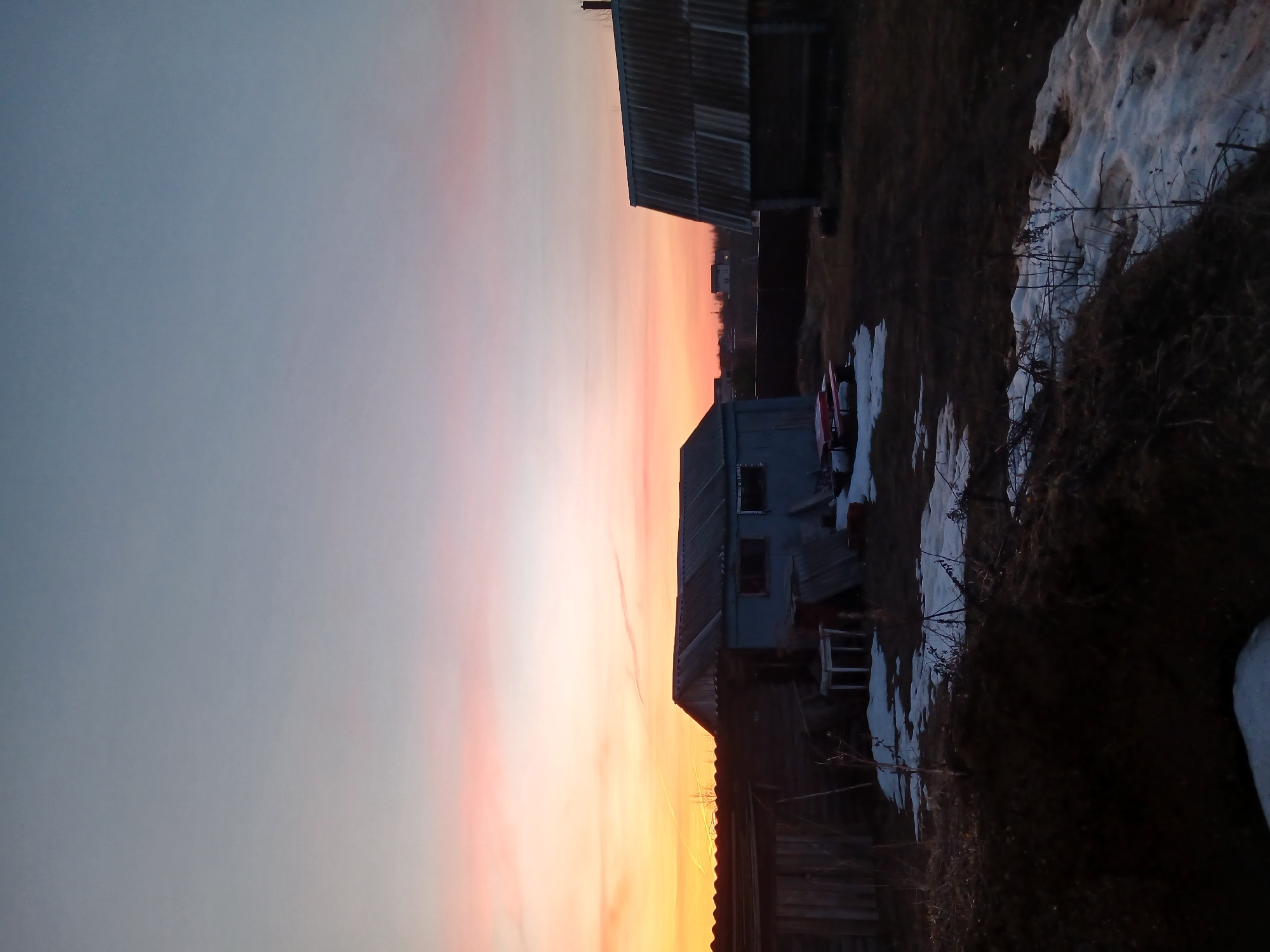 This landscape photograph captures a tranquil rural scene in Bolshaya Sosnova, Russia, likely during either sunset or sunrise. The image is composed with a wide expanse of sky dominating the upper portion of the frame, displaying a striking gradient of colors. Starting from a deep, muted greyish-blue at the top, the sky transitions smoothly to vibrant oranges and soft pinks closer to the horizon, indicative of the low angle of the sun. A faint, linear cloud formation or possibly a contrail is subtly visible across the colorful part of the sky.
In the foreground and mid-ground, several rustic buildings are silhouetted or partially illuminated against the bright sky. To the left, a large, dark building, possibly a barn or shed with a corrugated metal roof, stands prominently. Further to the right and slightly lower, another smaller structure, appearing to be a light-blue colored house or utility building with visible windows and a sloping roof, is present. The architectural style suggests typical village or countryside dwellings.
The ground beneath the structures is a blend of dark, dry earth and scattered patches of melting snow, indicating a cold season such as late winter or early spring. Sparse, twiggy vegetation and dried grasses protrude from the ground and snow. The scene is devoid of people or discernible activities, contributing to a serene and solitary atmosphere. The dramatic natural light contrasts with the subdued tones of the foreground elements, creating a peaceful and somewhat melancholic mood. No text is visible in the image.