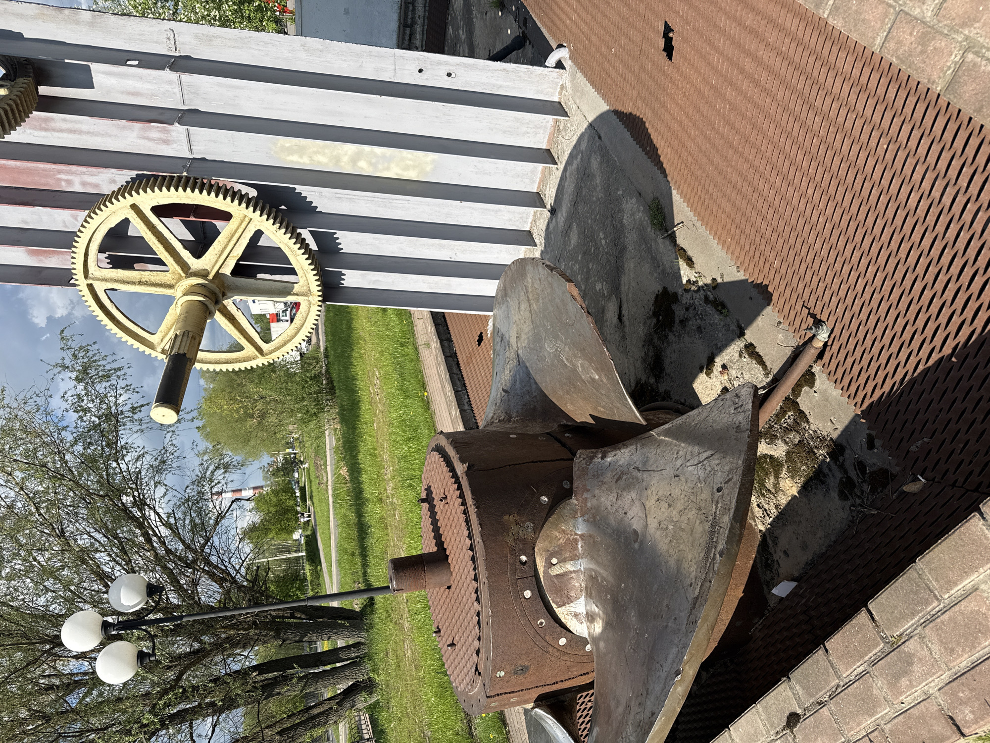 The image shows an outdoor display in Dmitrov, Russia, featuring decommissioned industrial machinery. Dominating the scene is a large, rusty, metal water turbine or pump, likely from a historical waterworks facility. It consists of a cylindrical housing, exposed blades, and protruding shafts. Next to it, mounted on a wall, is a large, yellow gear. The machinery is situated on a concrete base with brown metal grate and brick pavement. The surrounding environment includes a grassy area, trees, a street lamp, and in the background, other buildings and a smokestack. The image is taken during daylight hours, with the sky visible through tree branches.