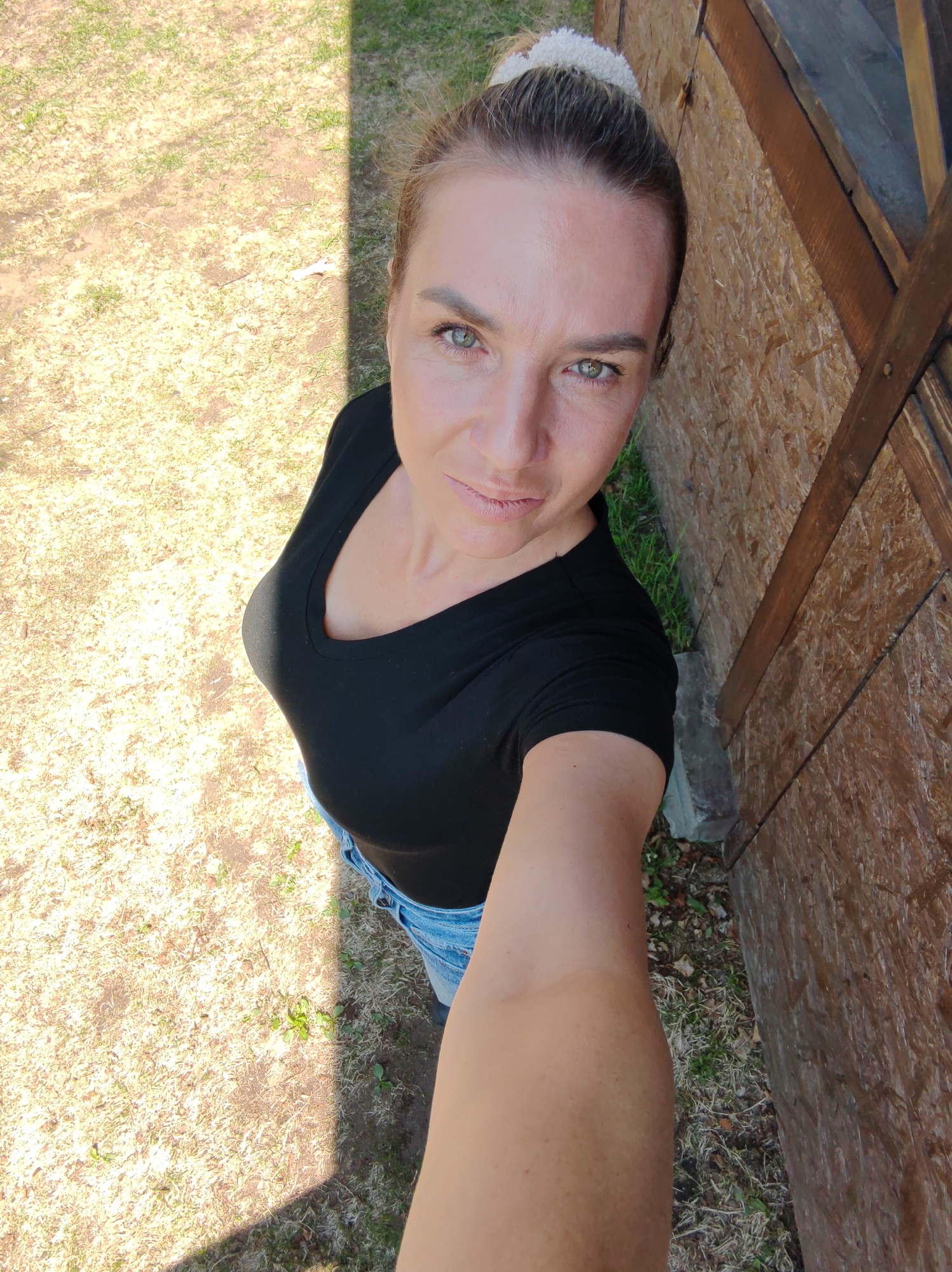 A forensic description of the scene in Unknown, Unknown:
A woman is captured in a high-angle selfie outdoors under bright sunlight. She is a light-skinned adult with light brown hair pulled into a bun secured by a white scrunchie. Her green eyes gaze directly into the camera, and her lips are lightly colored. She wears a black v-neck short-sleeved t-shirt and light blue denim jeans. Her right arm is visible, extended towards the camera, suggesting she is holding the device. The background to her left features dry, patchy grass and dirt, strongly illuminated by the sun, with a sharp diagonal shadow line bisecting the ground. To her right, a wooden structure, possibly a shed or fence, is visible. It appears to be constructed from oriented strand board (OSB) or similar pressed wood, with vertical and horizontal wooden supports. Sparse green grass grows at the base of this wooden structure.