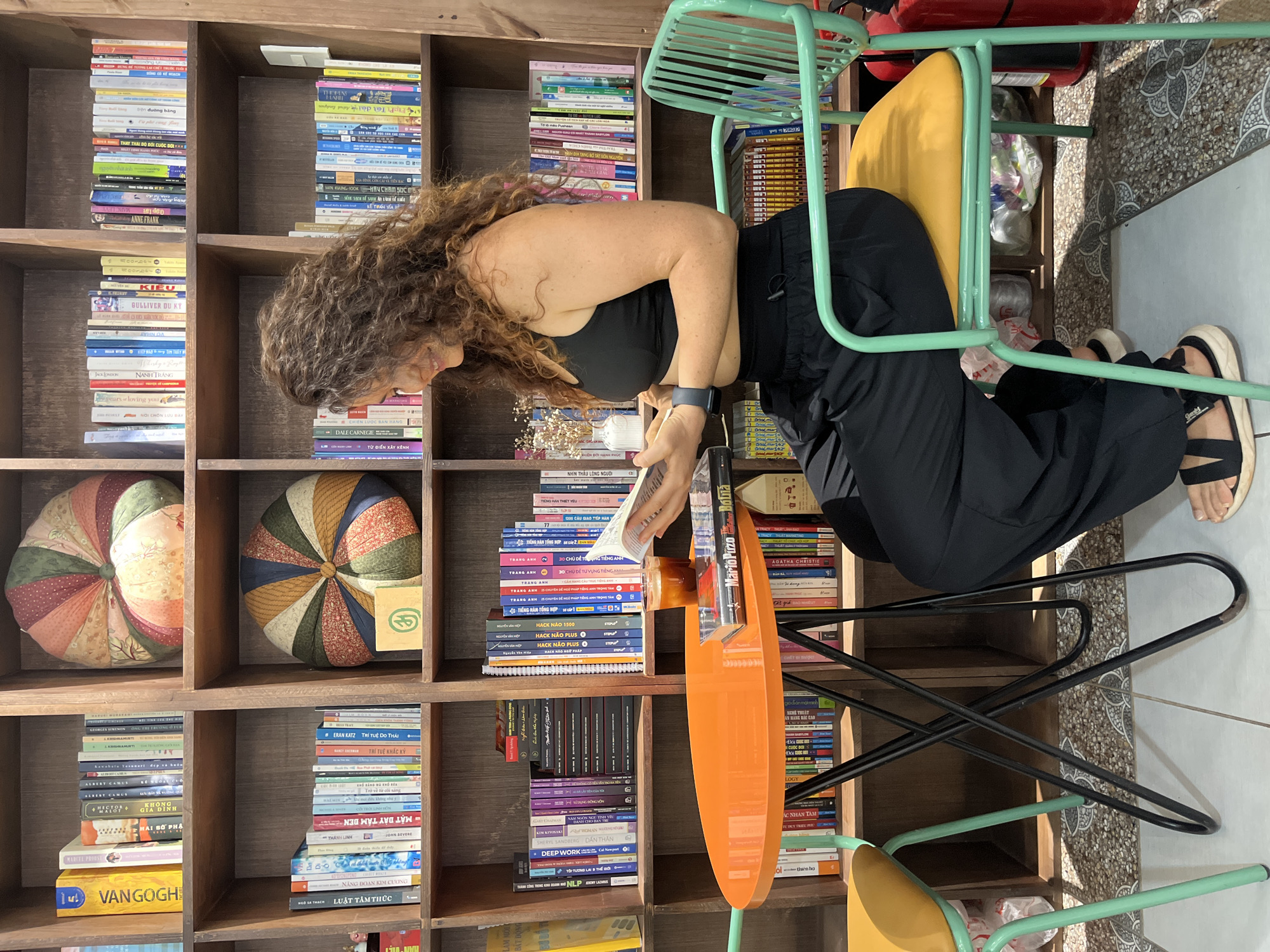 The image shows a woman sitting at an orange table in a library, reading a book. She is wearing a black sports bra and black pants. She has curly brown hair and is wearing sandals. On the table in front of her is a glass of iced coffee and two books. The library has shelves full of books and two round decorative pillows. The floor is tiled. The overall impression is one of a relaxed and cozy atmosphere.