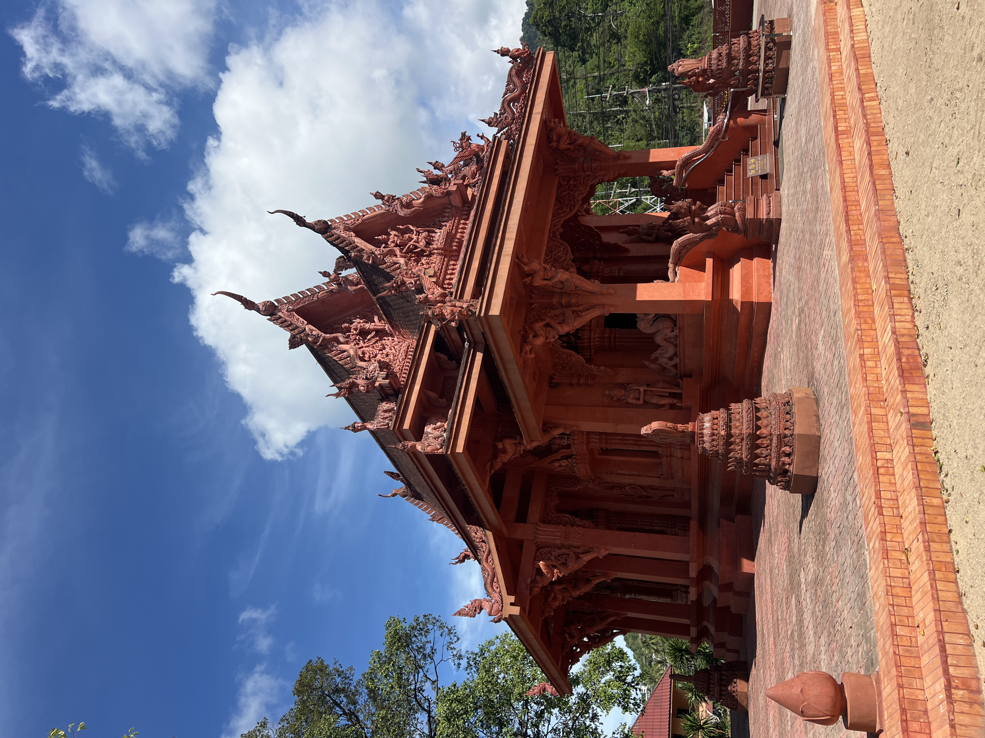 The image captures a vibrant red Buddhist temple in Baan Hua Thanon, Thailand. The temple's architecture is ornate, featuring multiple tiered roofs with intricate carvings of mythical creatures and figures. The building is constructed from red-colored material, possibly brick or plaster, which stands out against the clear blue sky dotted with white clouds.
In the foreground, a red tiled courtyard leads up to the temple entrance. Several decorative elements, including tiered stupas and a lotus-bud-shaped finial, are placed around the courtyard. The steps leading to the temple are also made of red tiles and are flanked by decorative railings and dragon sculptures.
To the left of the temple, a large green tree provides shade, with its leaves slightly blurred due to the angle. In the background, lush green hills rise, partially obscured by the temple's structure. The overall scene depicts a serene and culturally rich environment. There are no people visible in the image.