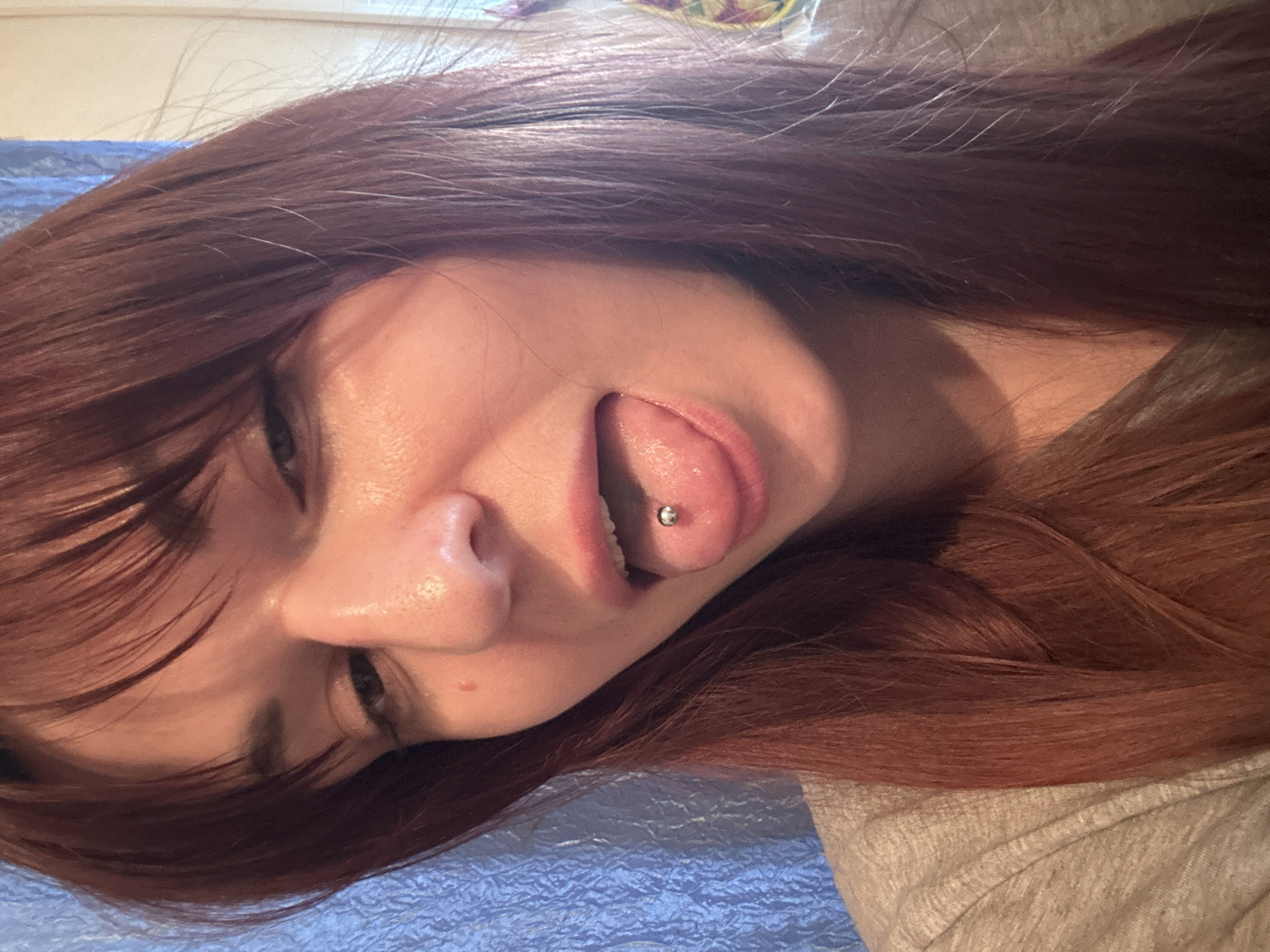 A medium shot depicts a female with red-auburn hair and fair skin lying on what appears to be a blue, textured surface, possibly a bed or blanket. The camera angle is tilted, framing her face from the lower left. She has a tongue piercing with a small silver ball and her tongue is partially extended. Her eyes are visible, but out of focus. The top right of the frame shows a portion of a light gray garment. The location is undetermined and there is no identifiable information to place the scene in Фоки, Russia.