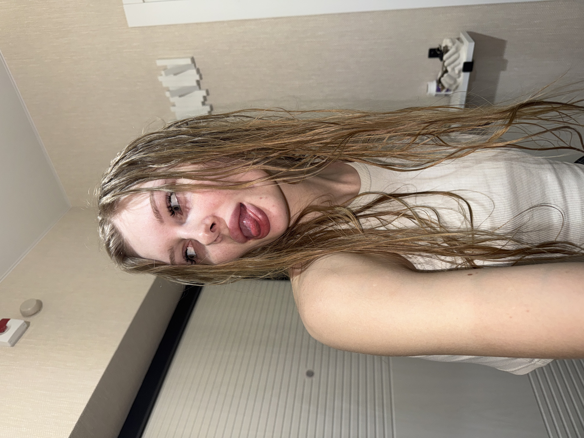 A young woman with blonde, wet hair is pictured in a selfie. She is wearing a white tank top and sticking her tongue out. The background appears to be a hotel room with a beige wall, a towel rack, and a door with vertical stripes. The location is unspecified, and there is no information that places it in Perm, Russia.