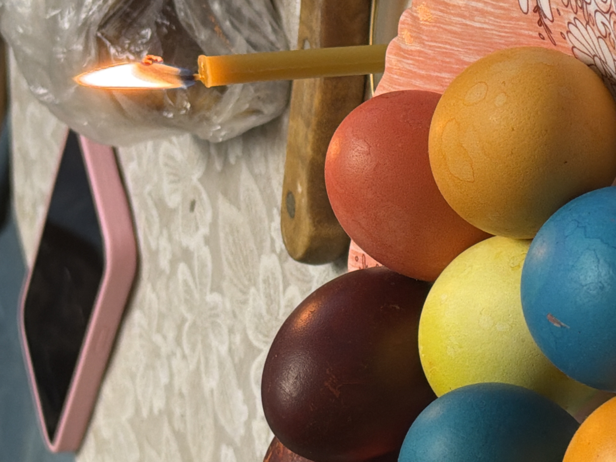A high-angle, close-up shot depicts a still life with several Easter eggs. The eggs are colored in shades of red, yellow, orange, and blue, and they are arranged in a haphazard stack. A burning candle lies horizontally across the top of the arrangement, with its flame pointing towards the left. The candle and eggs are situated next to a knife, which rests on a surface covered with a floral patterned tablecloth. To the left, a pink cellphone is visible on the tablecloth, and the background shows a plastic bag. The location may be Бронница, Russia.