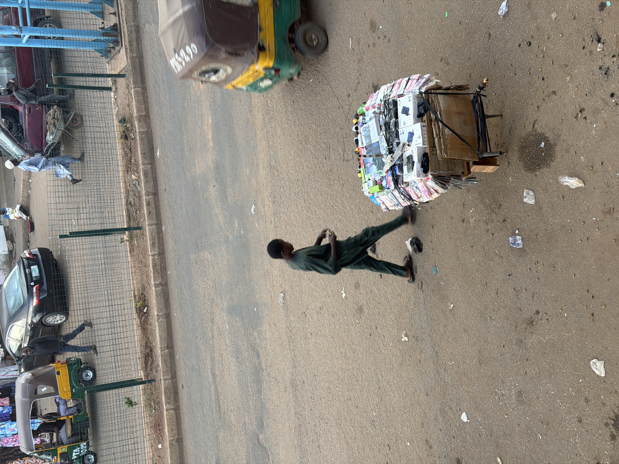 In Bukuru, Nigeria, an outdoor scene unfolds on a paved road surface with scattered debris. A young Black male, dressed in a dark green garment and sandals, walks towards the right of the frame. He is moving past a wooden cart laden with various items, including numerous phone cases and boxes, suggesting he might be a vendor or a passerby. To the right of the cart, a three-wheeled auto-rickshaw, predominantly yellow and green with a brown canopy, is visible. A Black male is seated in the driver's position of the auto-rickshaw, while another individual stands beside it. Further to the left, a black sedan is parked, and a Black male in a dark shirt and pants walks behind it. A green metal fence separates the road from a grassy area with a sidewalk. A portion of a burgundy colored vehicle is visible behind the fence, and another individual is seated nearby. Overhead, the sky is not visible.