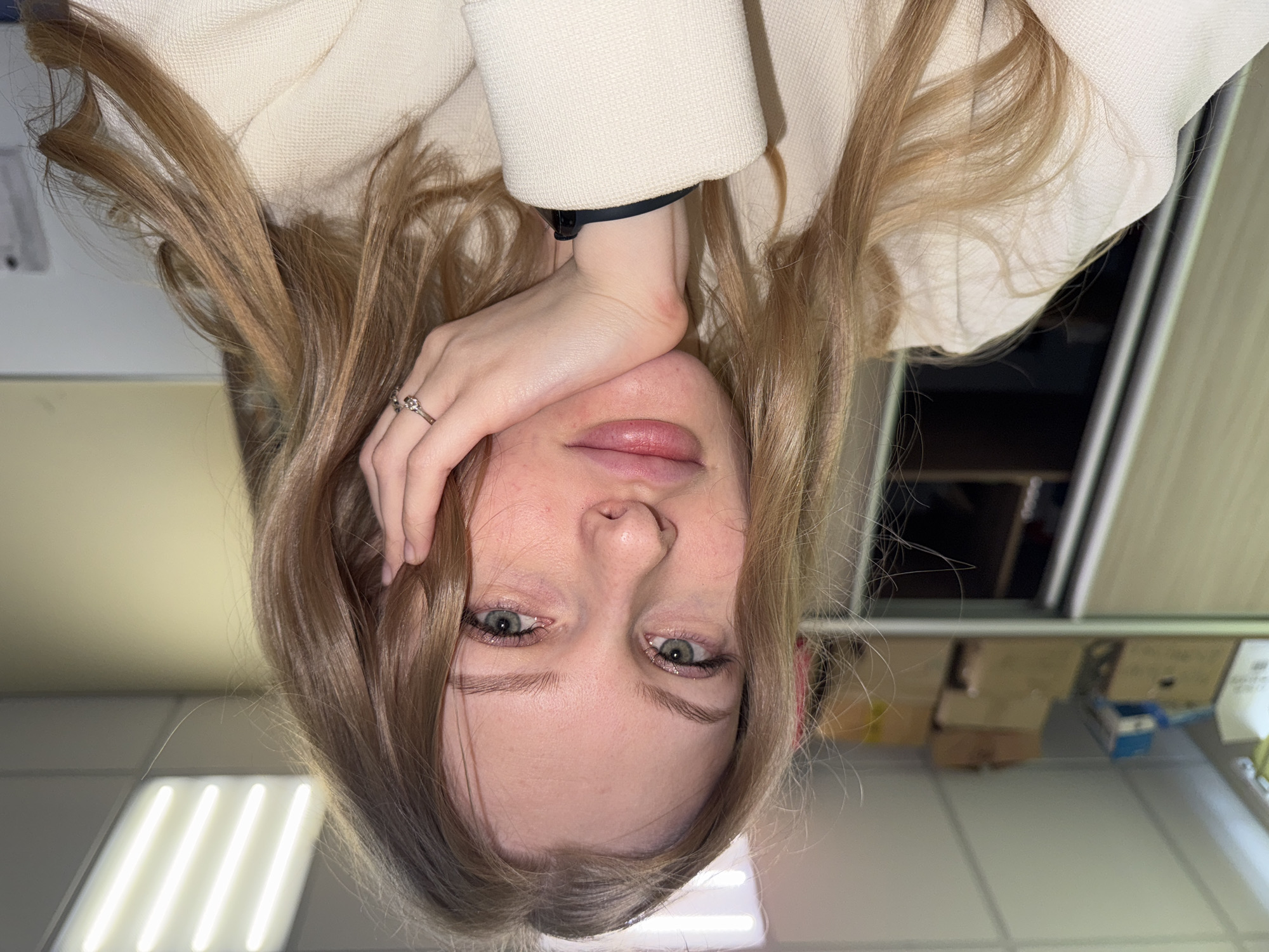 A person is upside down, with their face prominent in the frame. They have light brown hair and green eyes. Their hand is resting on their face, adorned with a ring on one finger. The person is wearing a light-colored jacket or coat. The background includes a shelf with boxes and supplies. Based on information available, the city of Perm, Russia has been noted for the location context.
