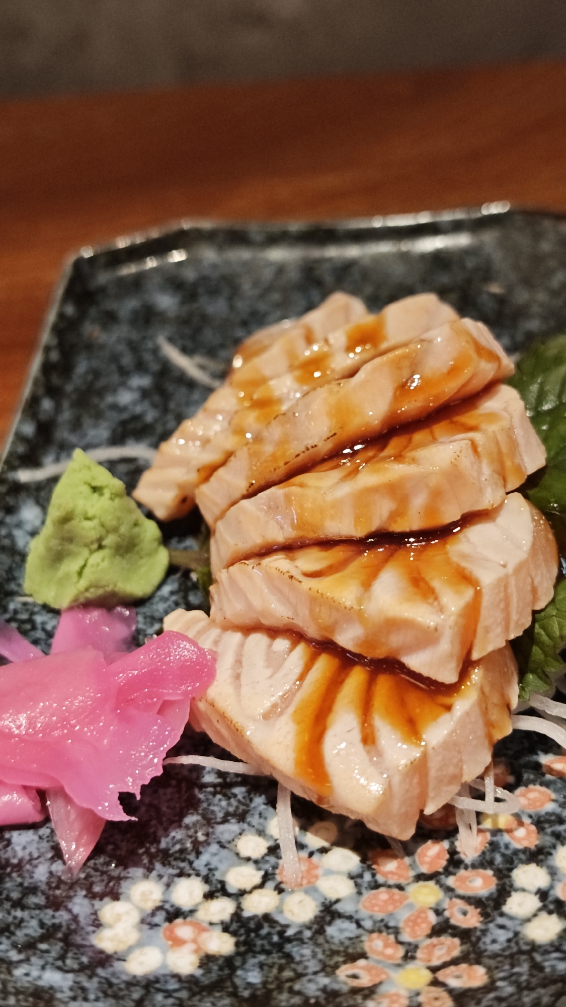 A close-up shot shows a plate of salmon sashimi in Đà Nẵng, Vietnam. The sashimi is neatly stacked, drizzled with a brown sauce, and accompanied by pickled ginger, wasabi, and shredded daikon radish. The plate itself is dark, patterned with flowers and dots. The background features a wooden surface.