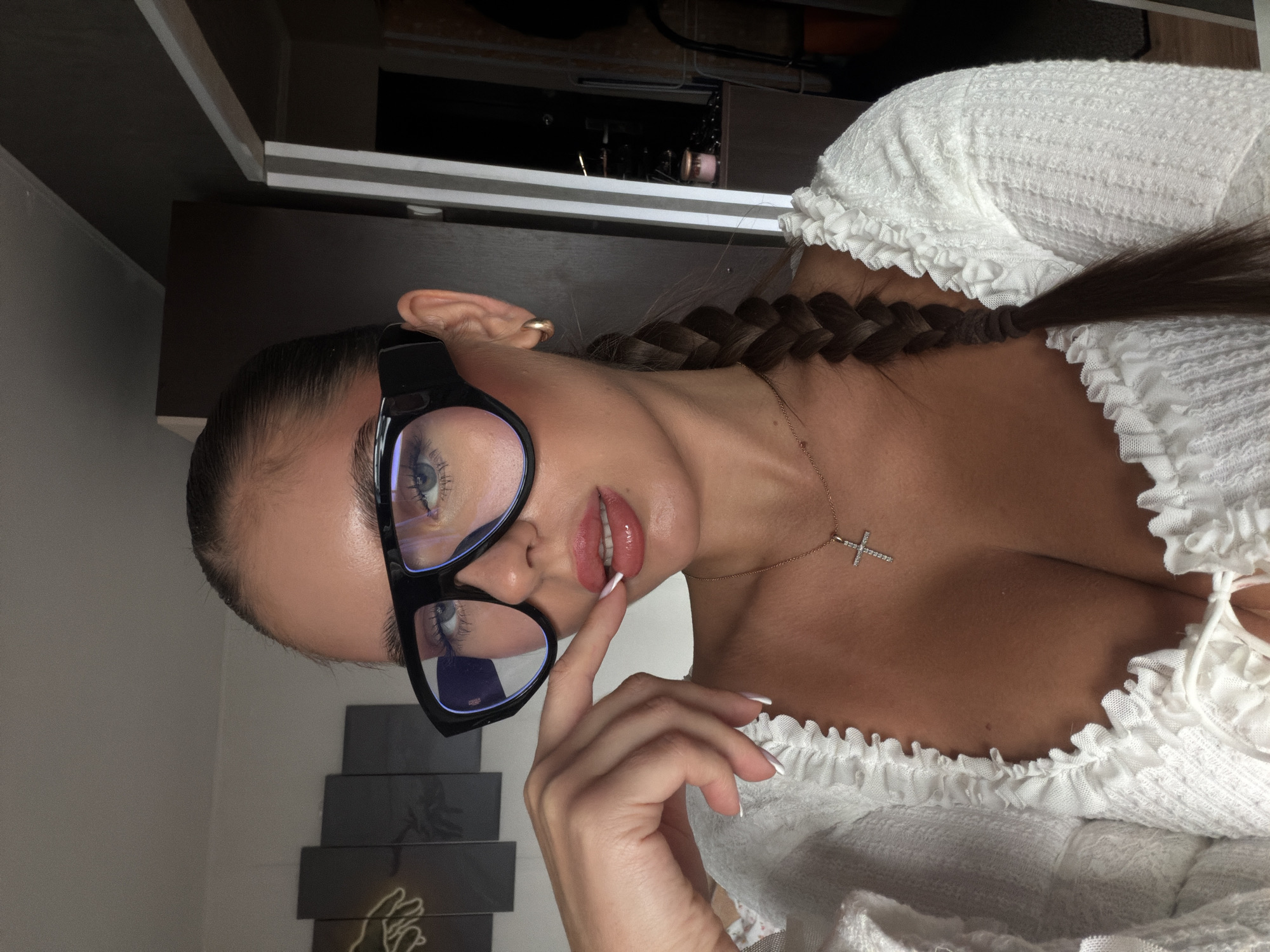 A woman is posing for a selfie. She has her hair braided, is wearing glasses and a white blouse, and has a cross necklace. The location cannot be determined from the image.