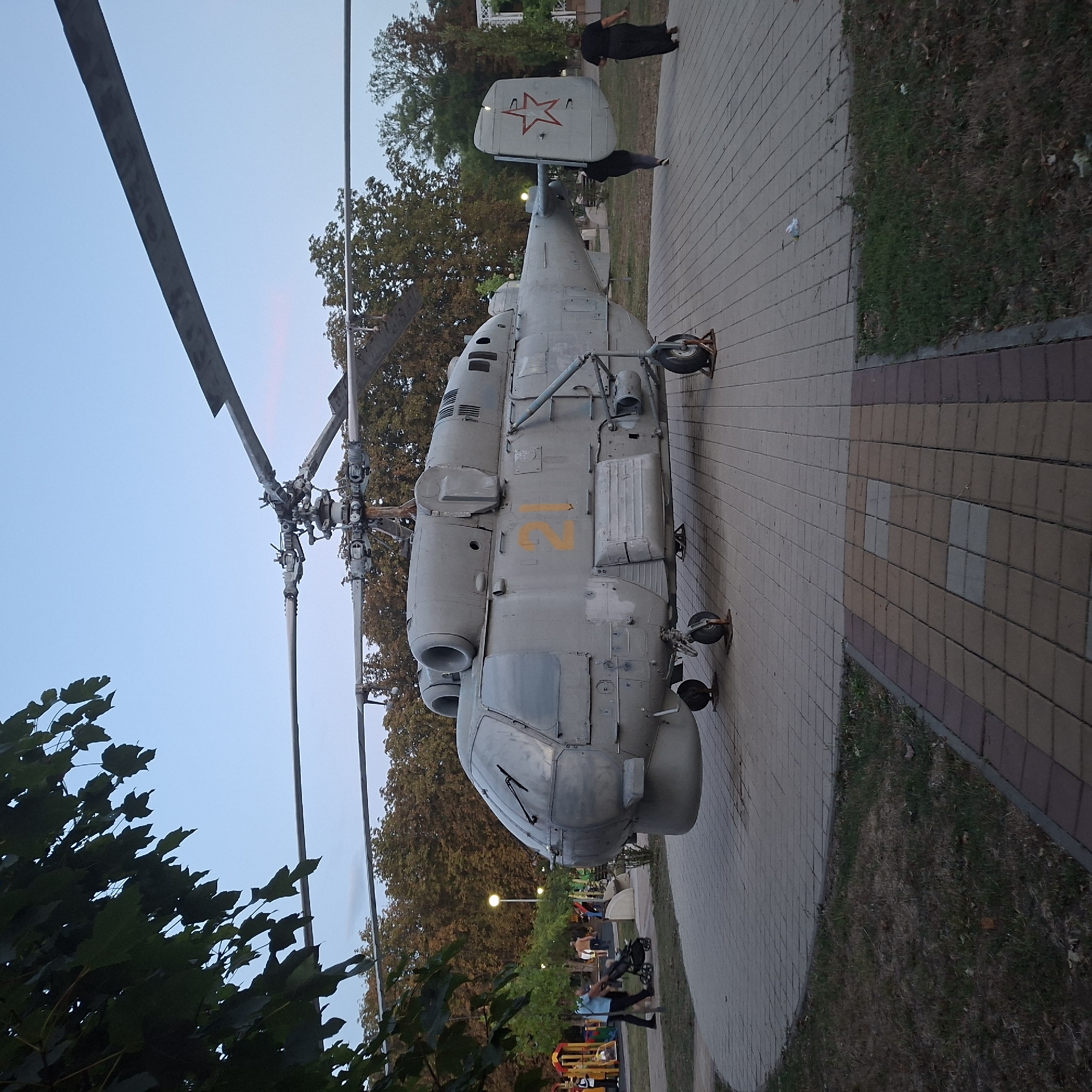 A large, grey helicopter, likely a monument or display, is parked on a paved circular area in what appears to be a public park. The helicopter has visible markings, including the number "21" in yellow on its side and a red star emblem on its tail fin. Two people are walking away from the helicopter on the paved area, and another person is pushing a stroller in the background, near a playground. Trees with autumn foliage are visible behind the helicopter, and the sky is a clear, pale blue, suggesting it's either early morning or late evening. The ground around the paved area is covered in grass and fallen leaves. The setting appears to be Anapskaya, Russia, based on the provided information.