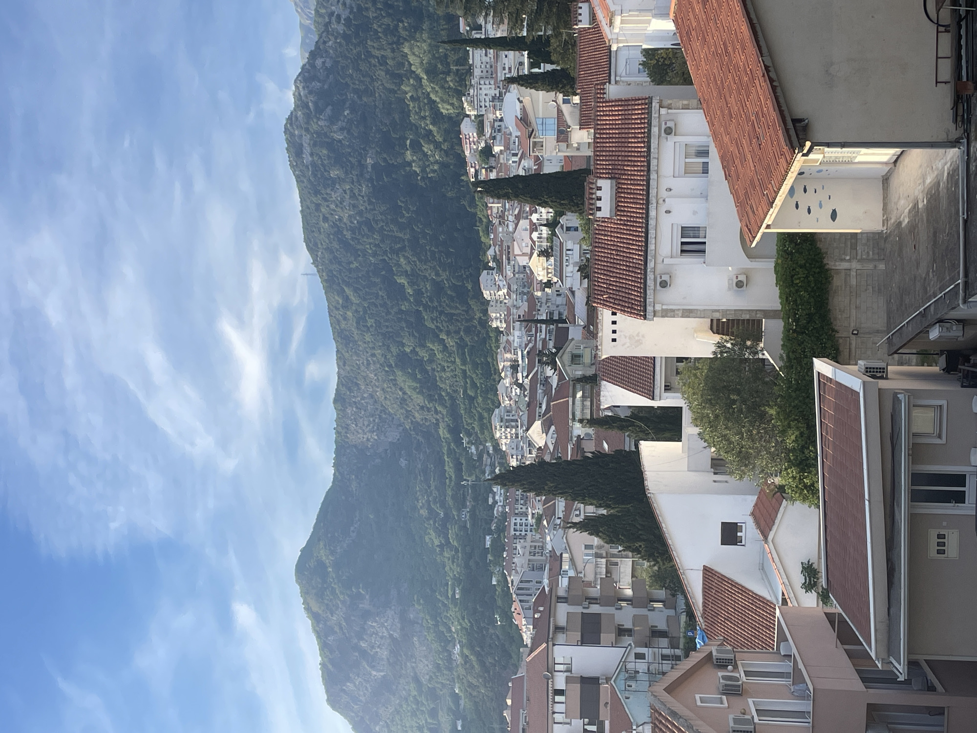 A high-angle, slightly tilted view captures a portion of Budva, Montenegro, showcasing a blend of natural and urban elements. A large, green-covered mountain dominates the left side of the frame, sloping down towards a dense arrangement of buildings. The buildings are primarily white with red-tiled roofs, characteristic of Mediterranean architecture. Cypress trees punctuate the landscape, adding vertical green accents amidst the built environment. The buildings are densely packed, suggesting a compact urban layout. The sky features a mix of light blue and scattered clouds. The scene appears to be shot from a building overlooking the city, with portions of rooftops and balconies visible in the foreground and right side of the image. No people are visible. The overall impression is one of a city nestled between the mountains and the Adriatic coast.