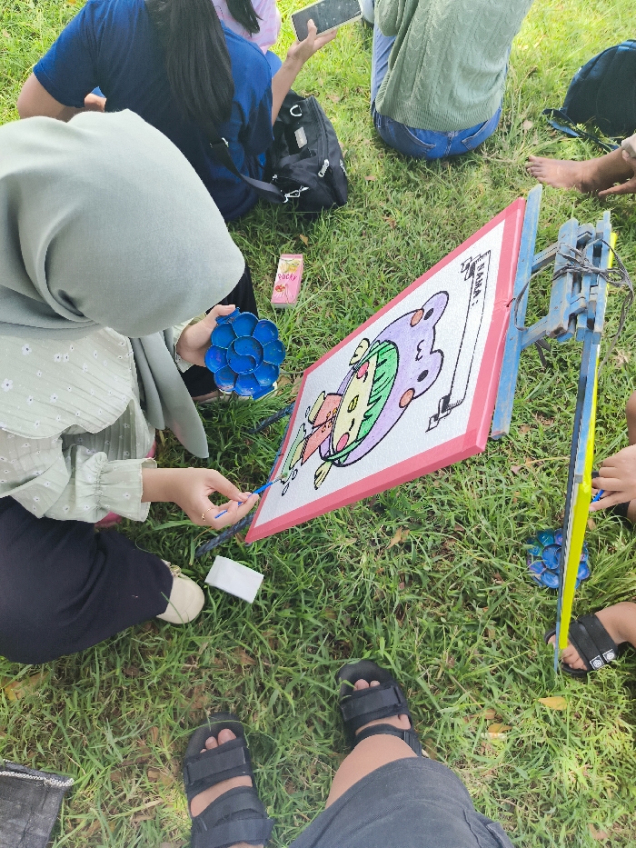 A woman wearing a light green headscarf and a floral patterned shirt paints a colorful cartoon character on a canvas easel. The scene is located in Majalengka, Indonesia, at the coordinates -6.846, 108.204. The woman is sitting on a grass lawn, holding a small blue paint tray and a blue brush. The easel is set up on the grass in front of her, with a bright pink, yellow, and green canvas attached. The canvas features a cartoon character with purple hair and a white shirt. The legs and feet of a second person are visible in the foreground, wearing black sandals. The background includes two other people, one sitting on the grass, wearing a green shirt, and the other sitting on the grass, wearing a blue shirt. There is a blue and black backpack lying in the grass nearby.