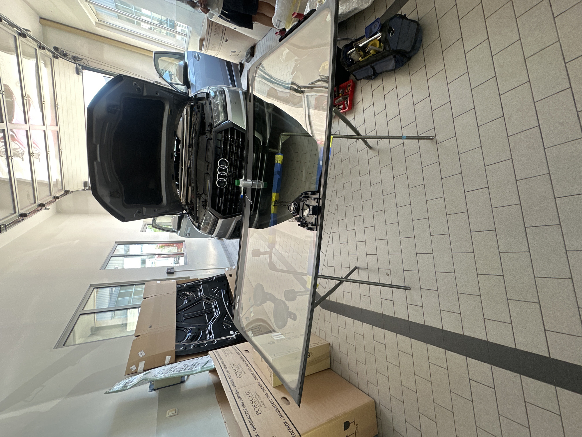An Audi SUV with its hood open is parked inside a garage with tiled flooring. In the foreground, a windshield is laid flat on a portable stand. Several cardboard boxes, one labeled "Porsche," are stacked to the left of the windshield. A black car hood is leaning against the boxes. To the right of the windshield, a blue tool bag and a red toolbox are on the floor. The garage has large windows and a large overhead door that is partially open, revealing the sky. The scene appears to be a car repair shop.