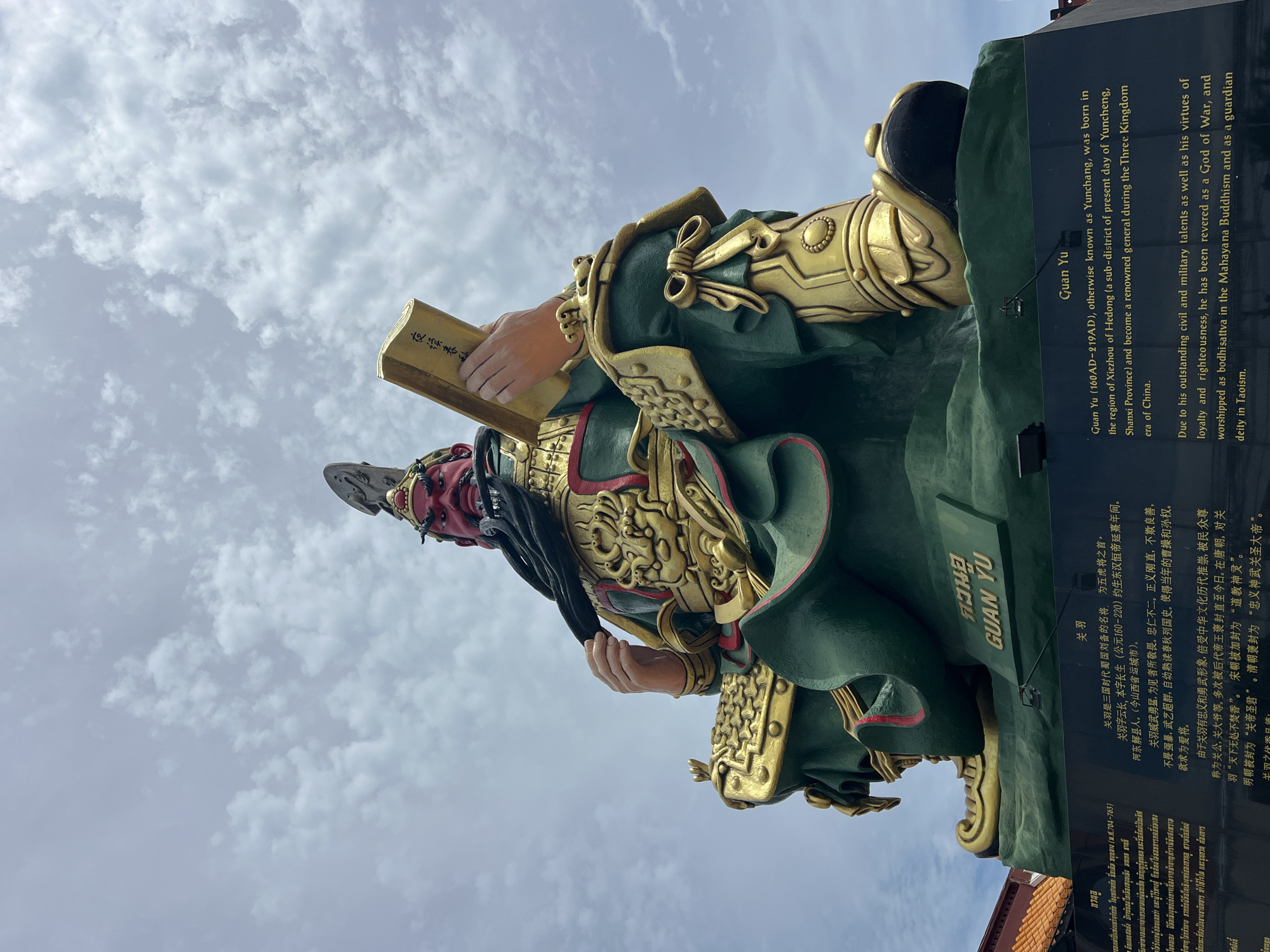 This is a medium-shot, eye-level photograph of a large, green and gold statue of Guan Yu, a deified Chinese general. The statue is located in the city of Baan Hua Thanon, Thailand. Guan Yu is depicted with a red face, black beard, and elaborate golden armor. He is holding a golden book in his right hand and his left hand is resting on his beard. The statue sits on a black, multi-leveled pedestal. Below the statue, on the black pedestal, is an inscription in both Thai and English. The English inscription reads: "Guan Yu (160AD-219AD), otherwise known as Yunchang, was born in the region of Xiezhou of Hedong (a sub-district of present day of Yuncheng, Shanxi Province) and become a renowned general during the Three Kingdom era of China. Due to his outstanding civil and military talents as well as his virtues of loyalty and righteousness, he has been revered as a God of War, and worshipped as bodhisattva in the Mahayana Buddhism and as a guardian deity in Taoism." The sky behind the statue is cloudy and overcast. To the left of the statue, a portion of a reddish-orange tiled roof is visible.